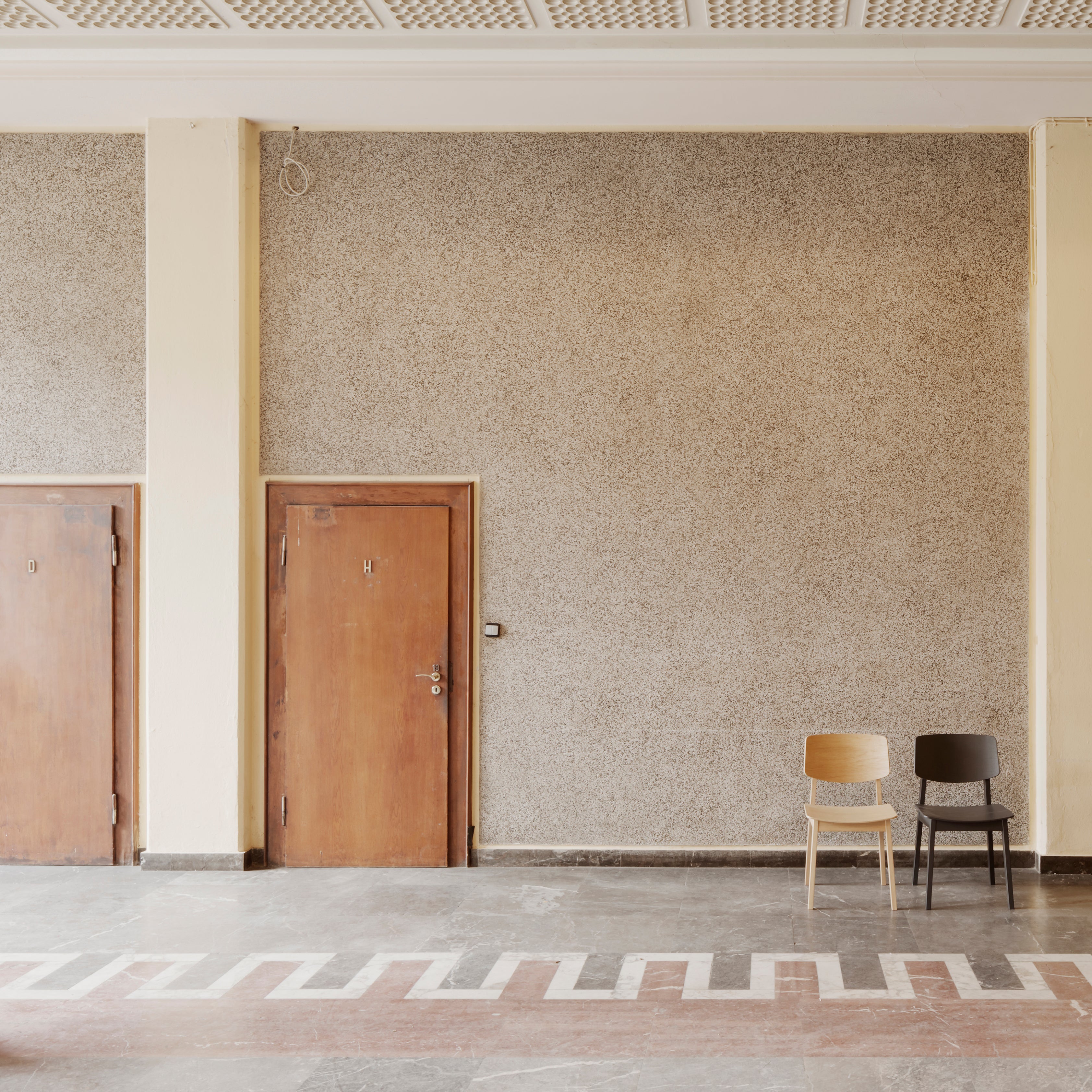 Two Usus Chairs by Bartmann Berlin, one in light oak and the other in dark oak, bring natural elegance to a spacious room with a textured wall featuring two closed wooden doors. Zigzag patterns enhance the flooring, and an intricate grid design adorns the ceiling.