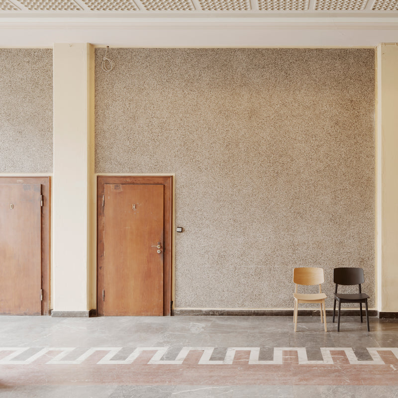 Two Usus Chairs by Bartmann Berlin, one in light oak and the other in dark oak, bring natural elegance to a spacious room with a textured wall featuring two closed wooden doors. Zigzag patterns enhance the flooring, and an intricate grid design adorns the ceiling.