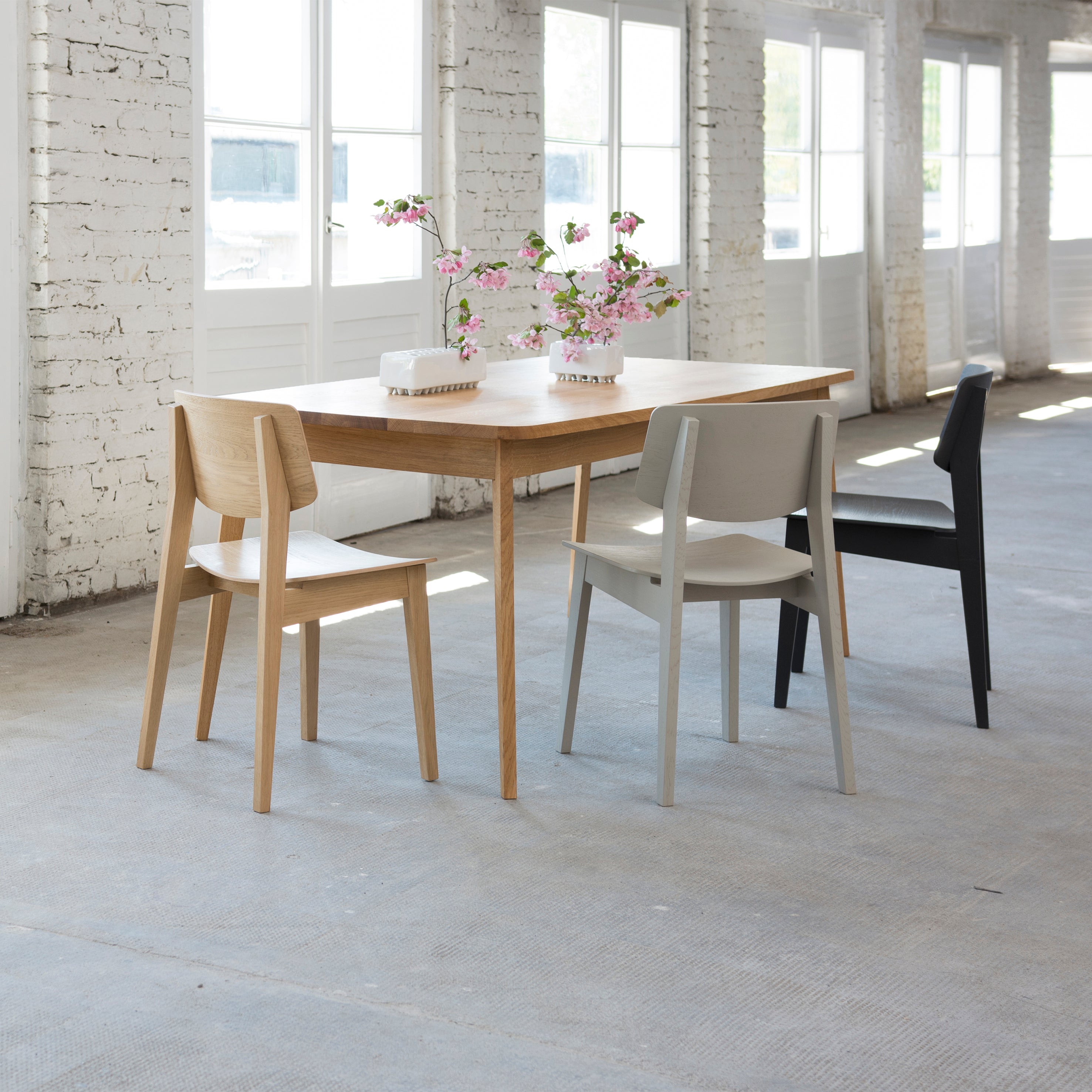 A minimalist dining area showcases the Bartmann Berlin Usus Table surrounded by three chairs—one wooden, one gray, and one black. Pink flowers in white vases decorate the table, while white brick walls and large windows illuminate the space.
