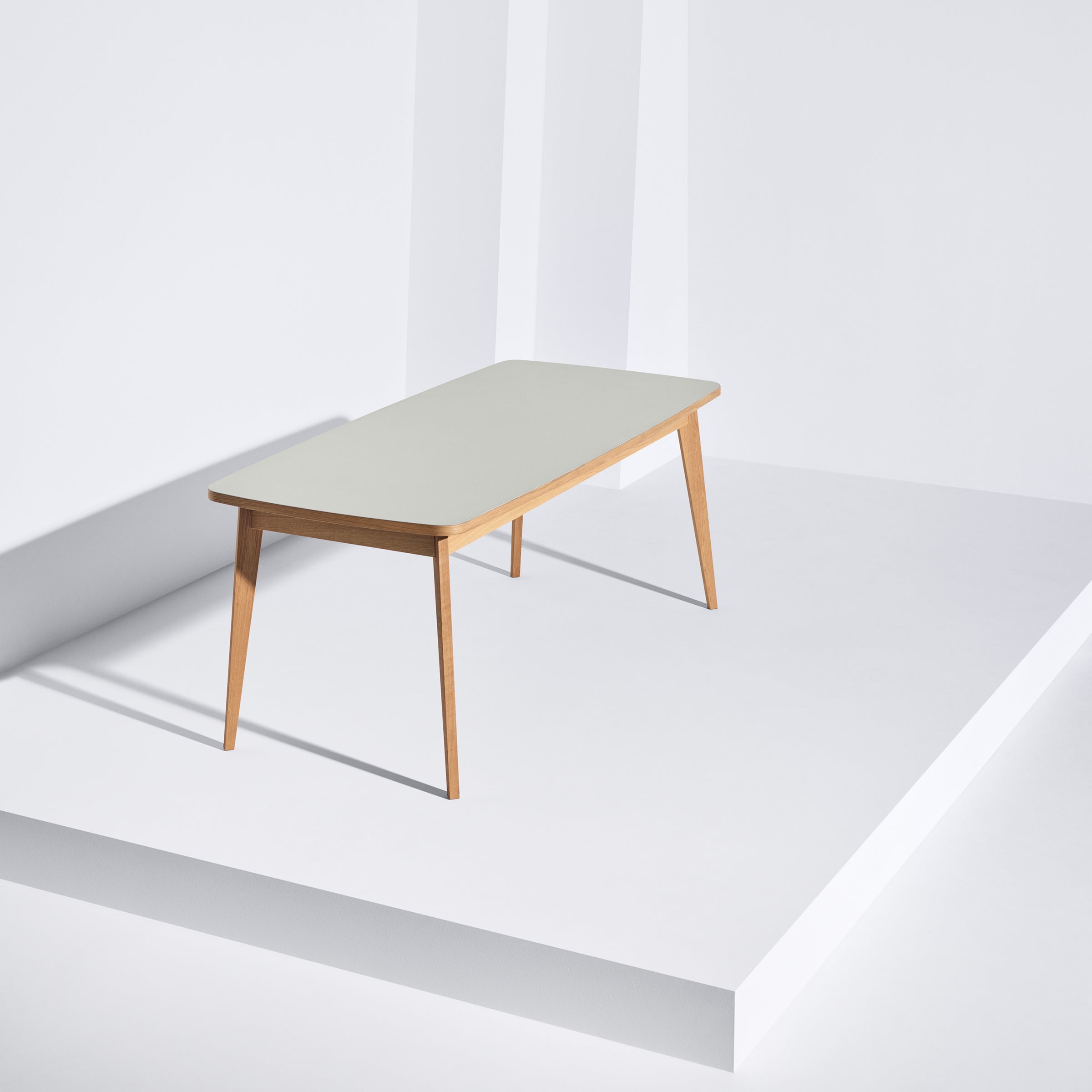 A minimalist Usus Table by Bartmann Berlin, crafted from European oak with a white linoleum-coated top and slender legs, is set on a white corner platform. Bright surroundings enhance the soft shadows against the walls.