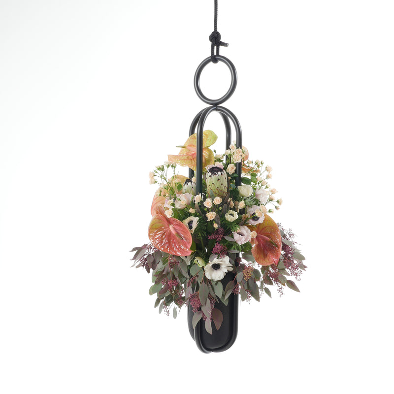 Atelier Haussmanns Blumenampel - Hanging Flower Vase showcases pink anthuriums, white flowers, and greenery in a black oval holder suspended by a black cord against a white background, echoing the refined style of the elegant Ashes II Vase.