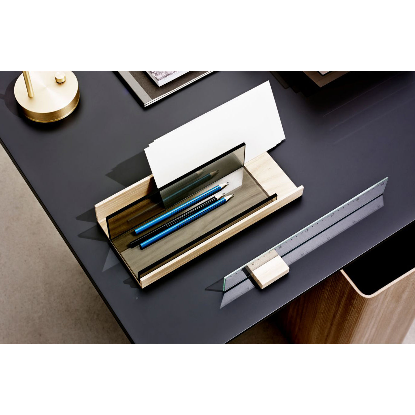 A modern office desk setup features Boewers Charlie Desk Accessories, with a ruler and transparent black stand holding white papers in Pauline Deltour design. A wooden holder with three blue pencils enhances the dark grey desk, alongside an elm and colored glass accessory.