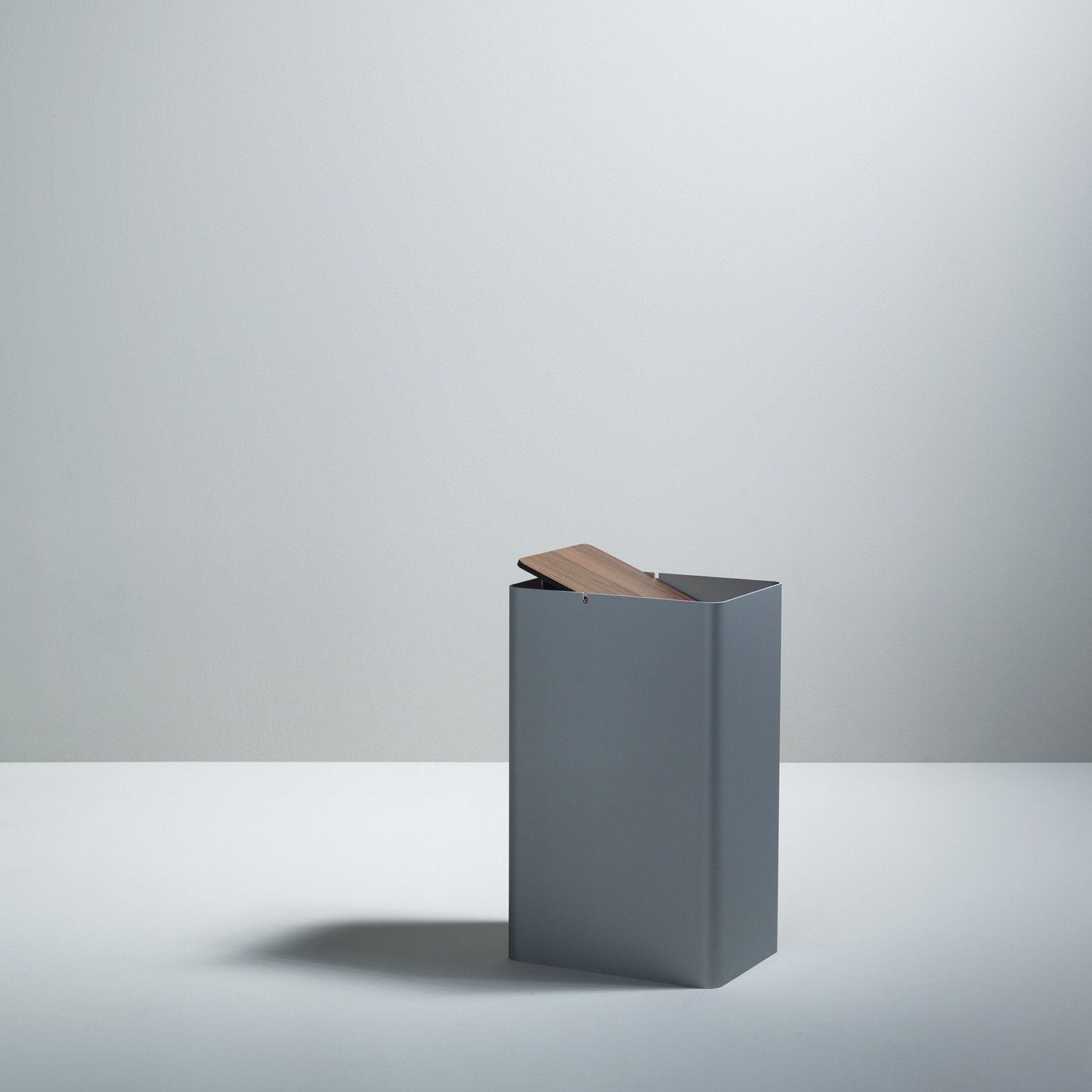 In a neutral-toned room, the Boewer P-3 minimalist square gray waste bin with a wooden lid is slightly open, casting a soft shadow on the light-colored floor.