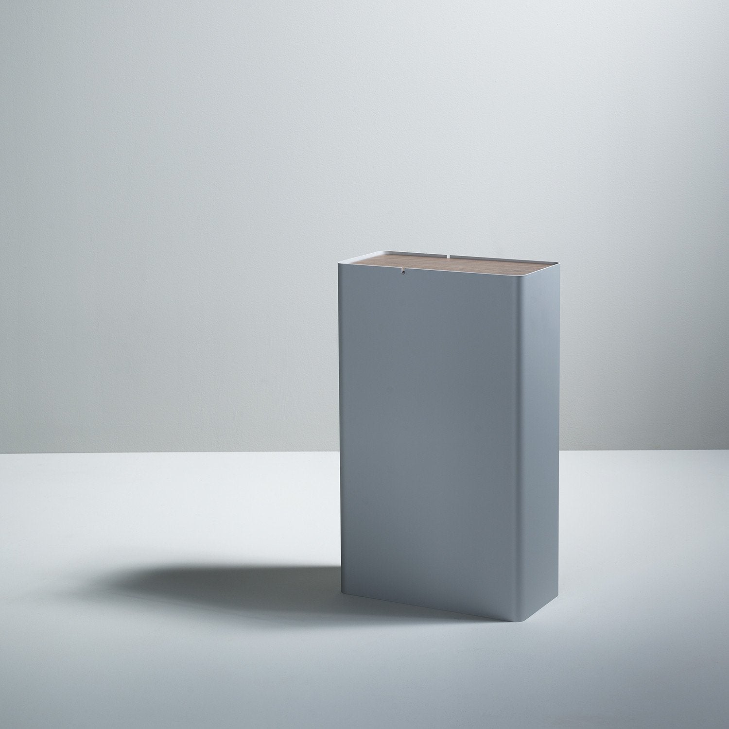 A Boewer P-3 minimalist gray waste bin with a closed lid stands against an off-white background, casting a subtle shadow on the floor. Its sleek design is reminiscent of modern aluminum containers.