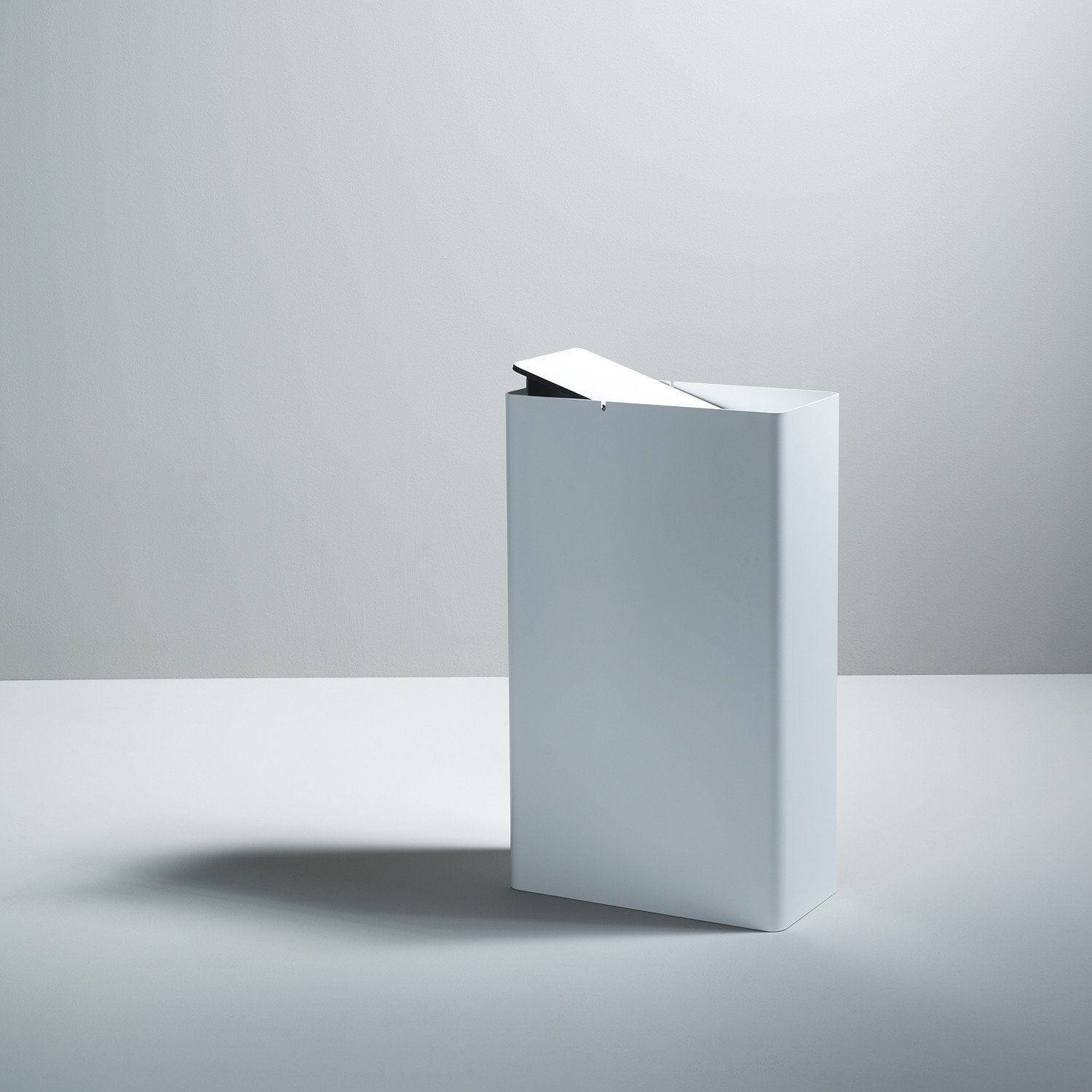 The Boewer P-3 is a minimalist white waste bin with a slightly open lid, placed on a plain surface against a light gray background. Its clean, modern design echoes sleek aluminum containers, casting subtle shadows to the left.