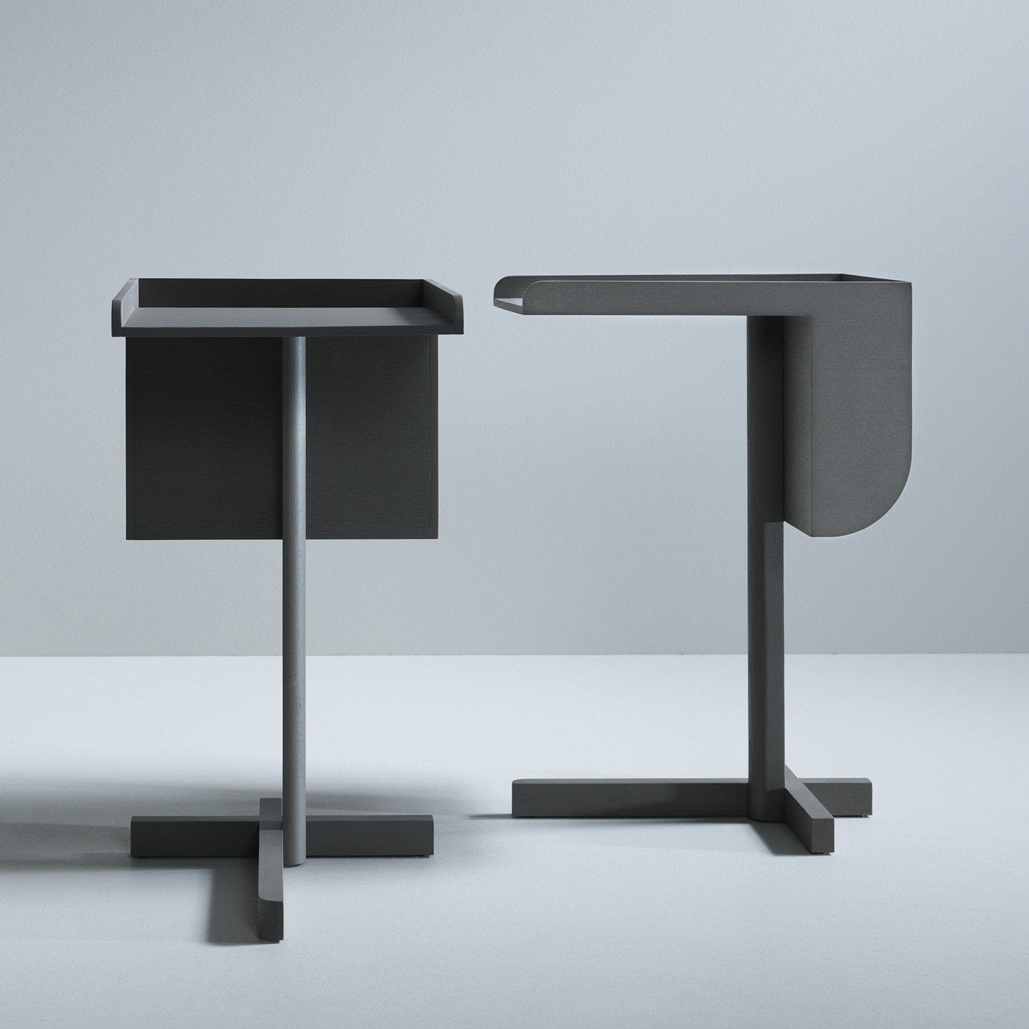 Two modern, minimalist side tables from Boewers School series, echoing Konstantin Grcics style, stand against a light gray background. Both are dark gray with unique geometric shapes and clean lines: one open design and the other enclosed.