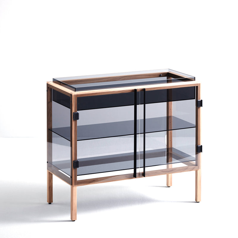 The Boewer Shade is a modern glass display cabinet with a solid elm frame and two sliding doors. It features adjustable shelves, stands on four legs, and its smoky-tinted glass complements the light wood finish against a plain white surface.