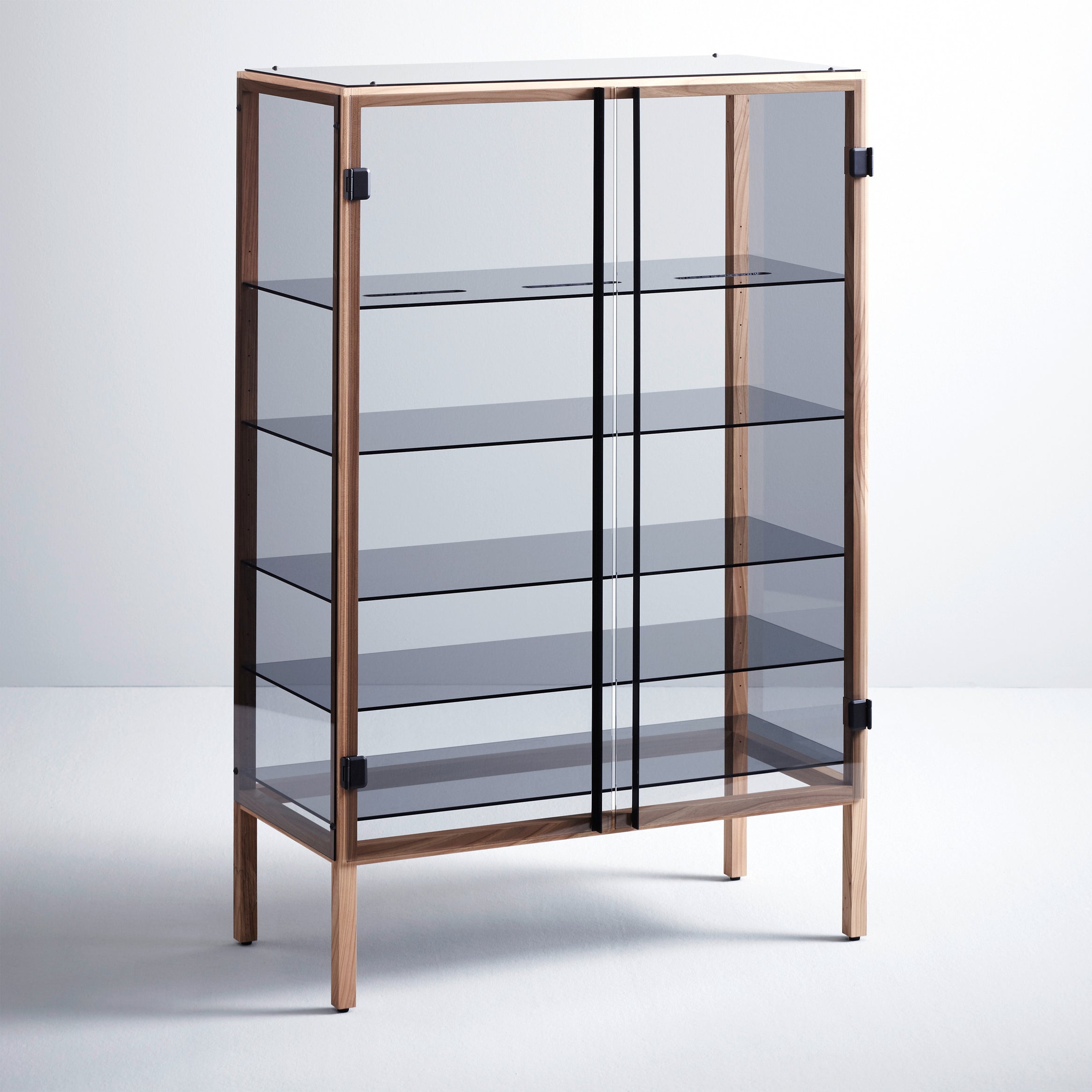 The Shade display cabinet by Boewer is a modern piece with transparent glass doors and sides, black metal accents, and wooden edges. It features four adjustable glass shelves and is supported by solid elm legs, perfectly complementing a plain white wall and light gray floor.