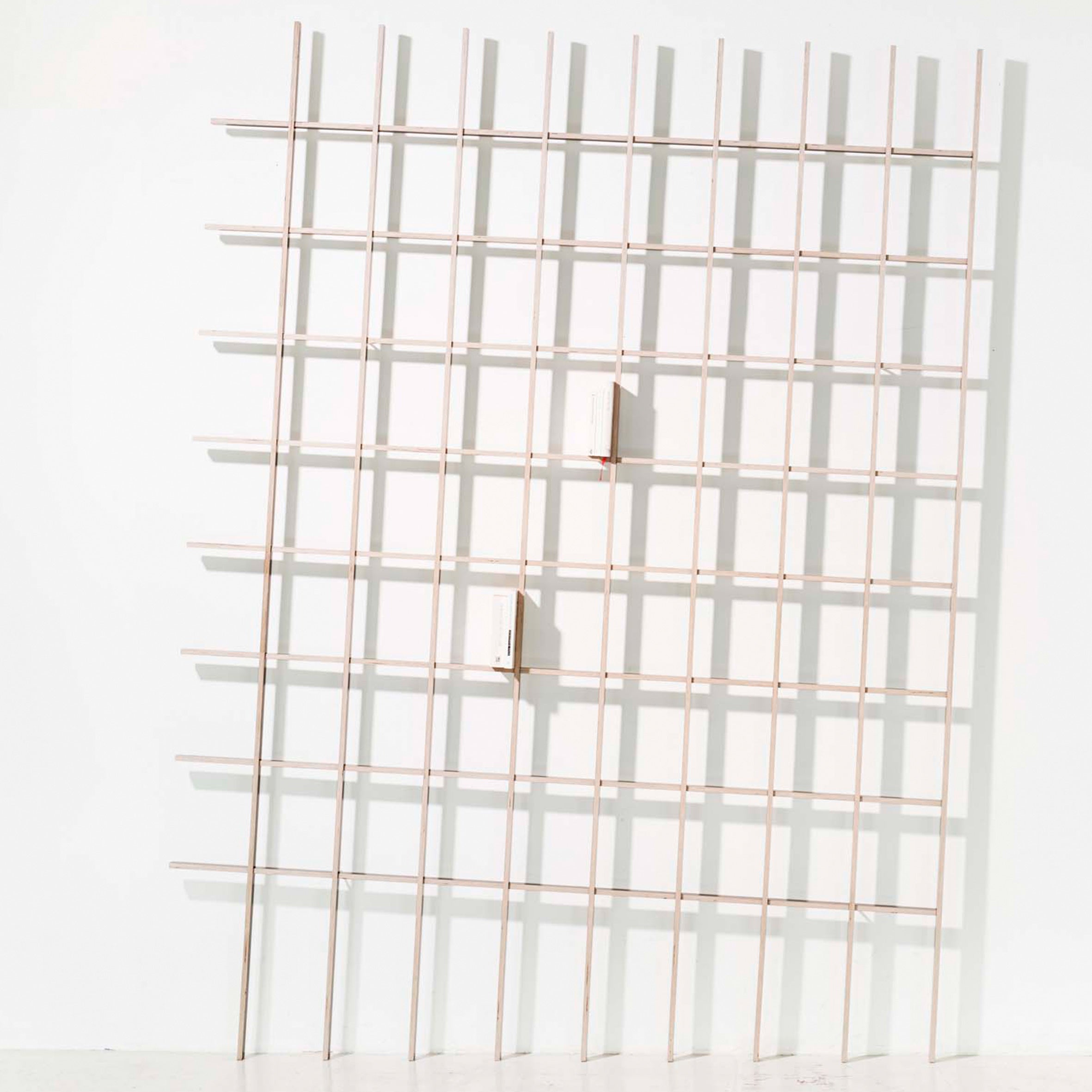 The SCHLAGSEITE by Martin Breuer Bono features a grid-like structure of thin wooden slats on a white wall. Two small white rectangular blocks are attached, creating depth and elegance in this minimalist furniture piece.