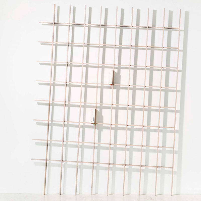 The SCHLAGSEITE by Martin Breuer Bono features a grid-like structure of thin wooden slats on a white wall. Two small white rectangular blocks are attached, creating depth and elegance in this minimalist furniture piece.