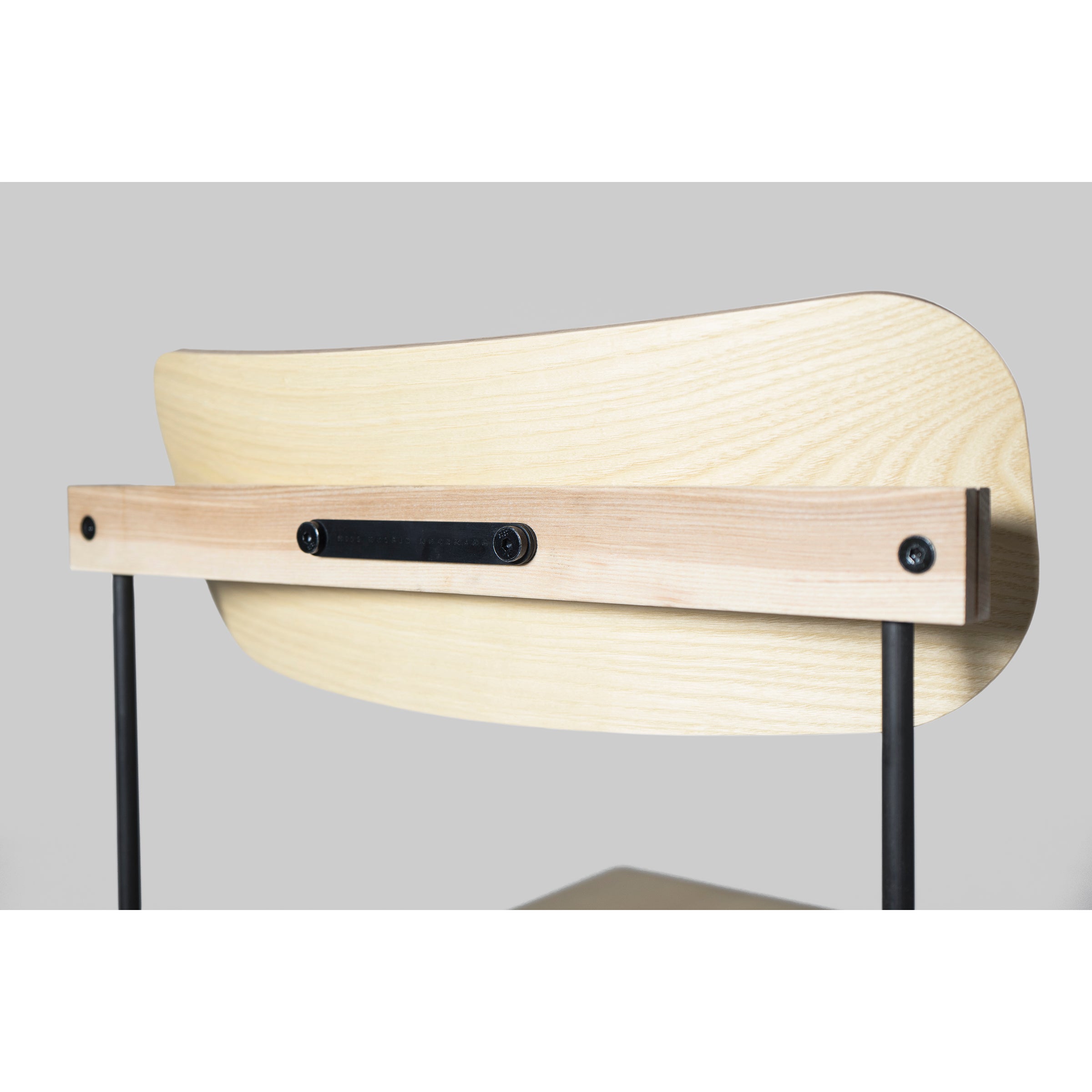 A close-up of the Bruto chair by Moormann displays its backrest, made from molded plywood with a light ash veneer. The horizontal metal bar, attached to two vertical black metal supports with screws, contrasts against a plain gray background.