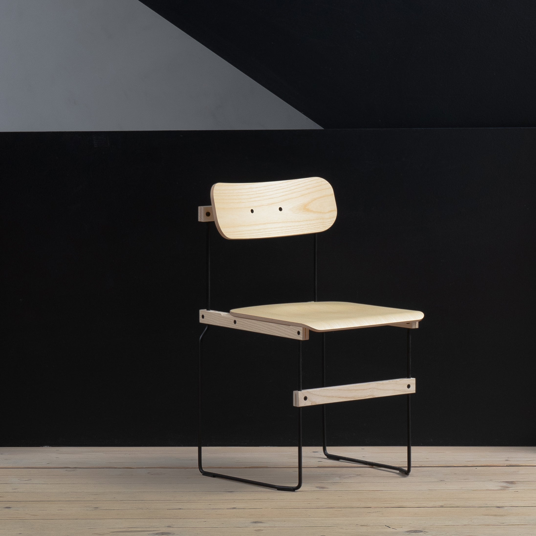 A Moormann Bruto chair, featuring a minimalist ash veneer finish and a black metal frame, is set against a dark wall and on a light wooden floor, showcasing its simple, modern design with clean lines and functionality.