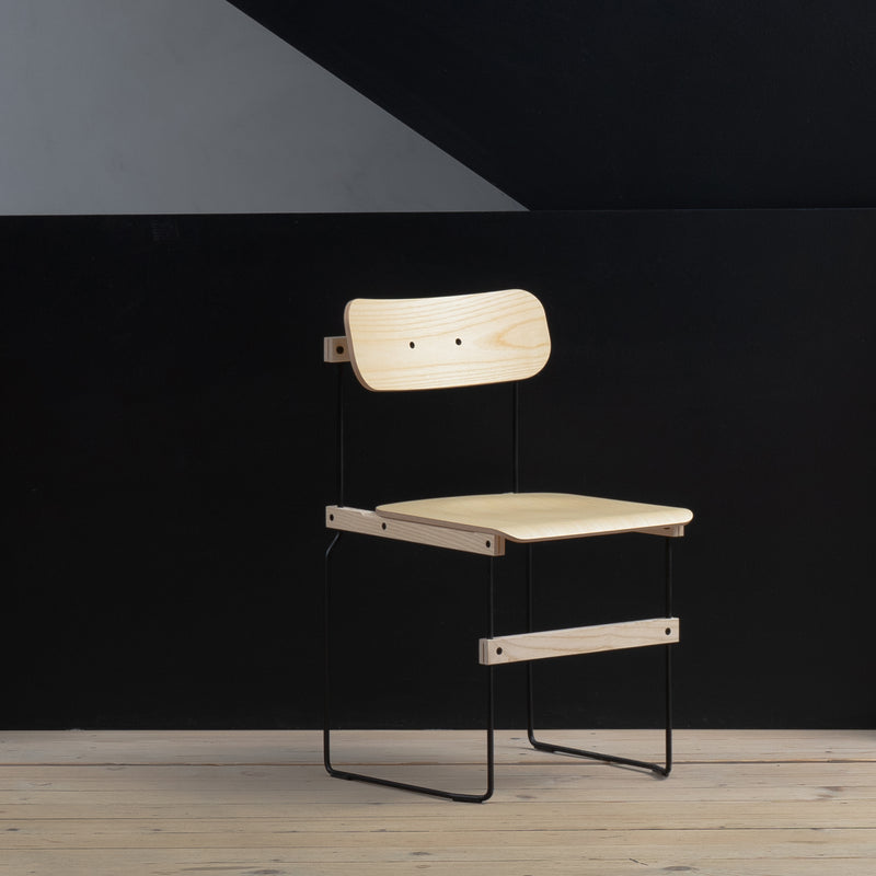 A Moormann Bruto chair, featuring a minimalist ash veneer finish and a black metal frame, is set against a dark wall and on a light wooden floor, showcasing its simple, modern design with clean lines and functionality.