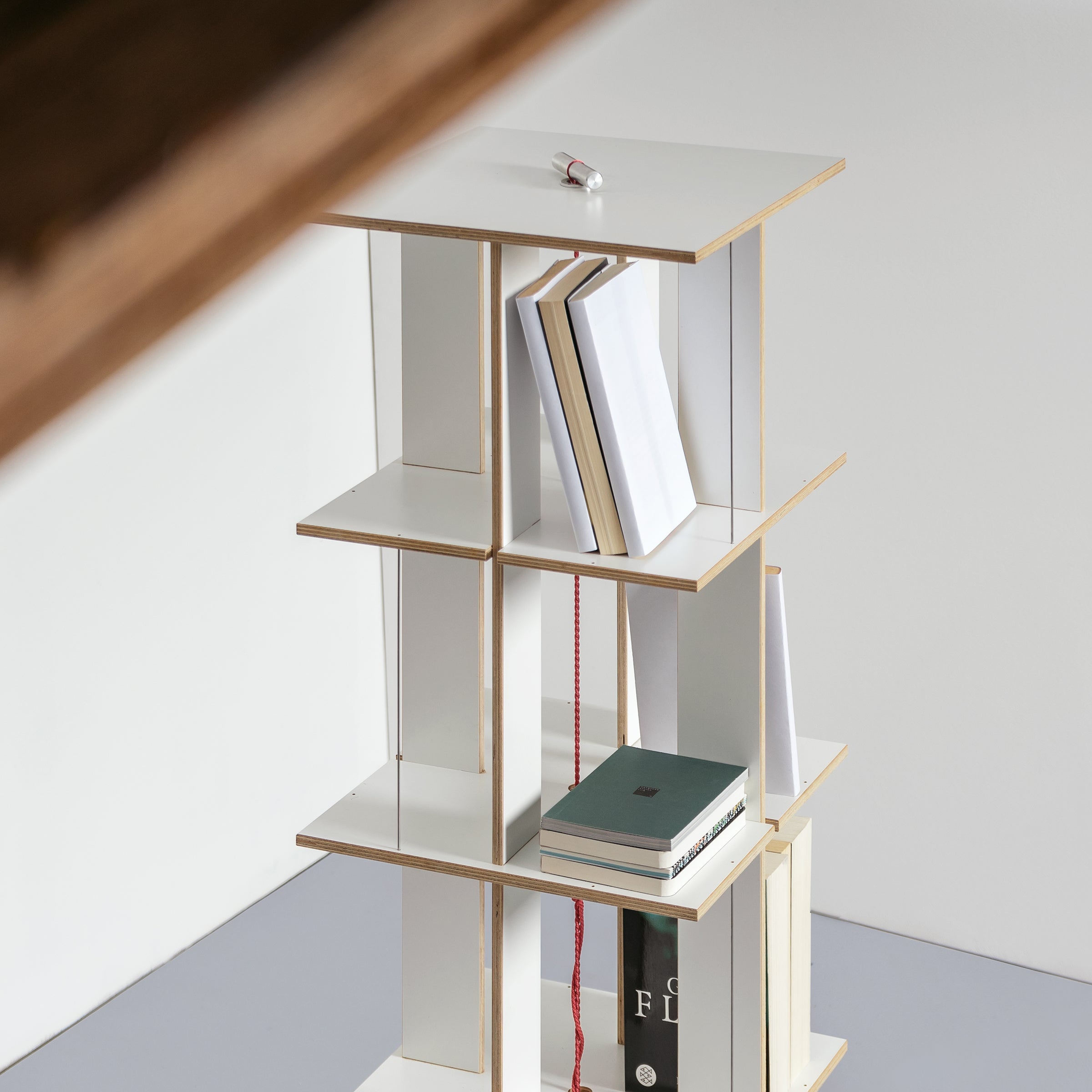 The Moormann Buchstabler rack has staggered white shelves that provide space-saving storage with a geometric design, complemented by a small item on top and a simple background to highlight its modern aesthetic.