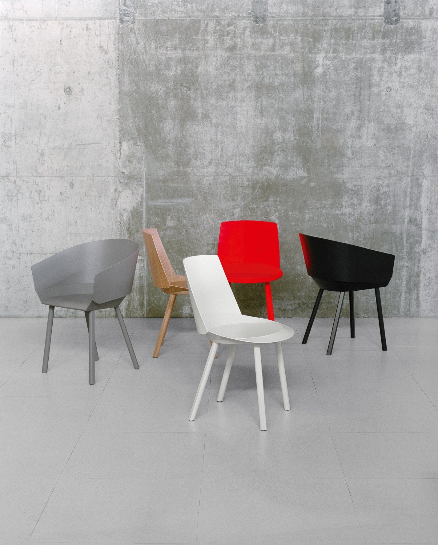 A set of five CH05 THIS Side Chairs by E15, crafted from oak veneer plywood in minimalist colors—gray, wood, red, black, and white—is elegantly displayed on a light gray floor against a textured concrete wall.