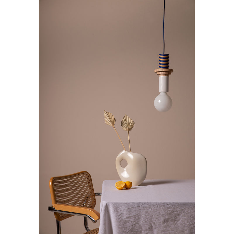 A minimalist room showcases a wicker chair, an ash wood table covered by a white cloth, and a white vase with two dried leaves. A lemon slice adorns the table while a Schneid Junit Column light bulb hangs above, casting its glow against the serene beige wall.