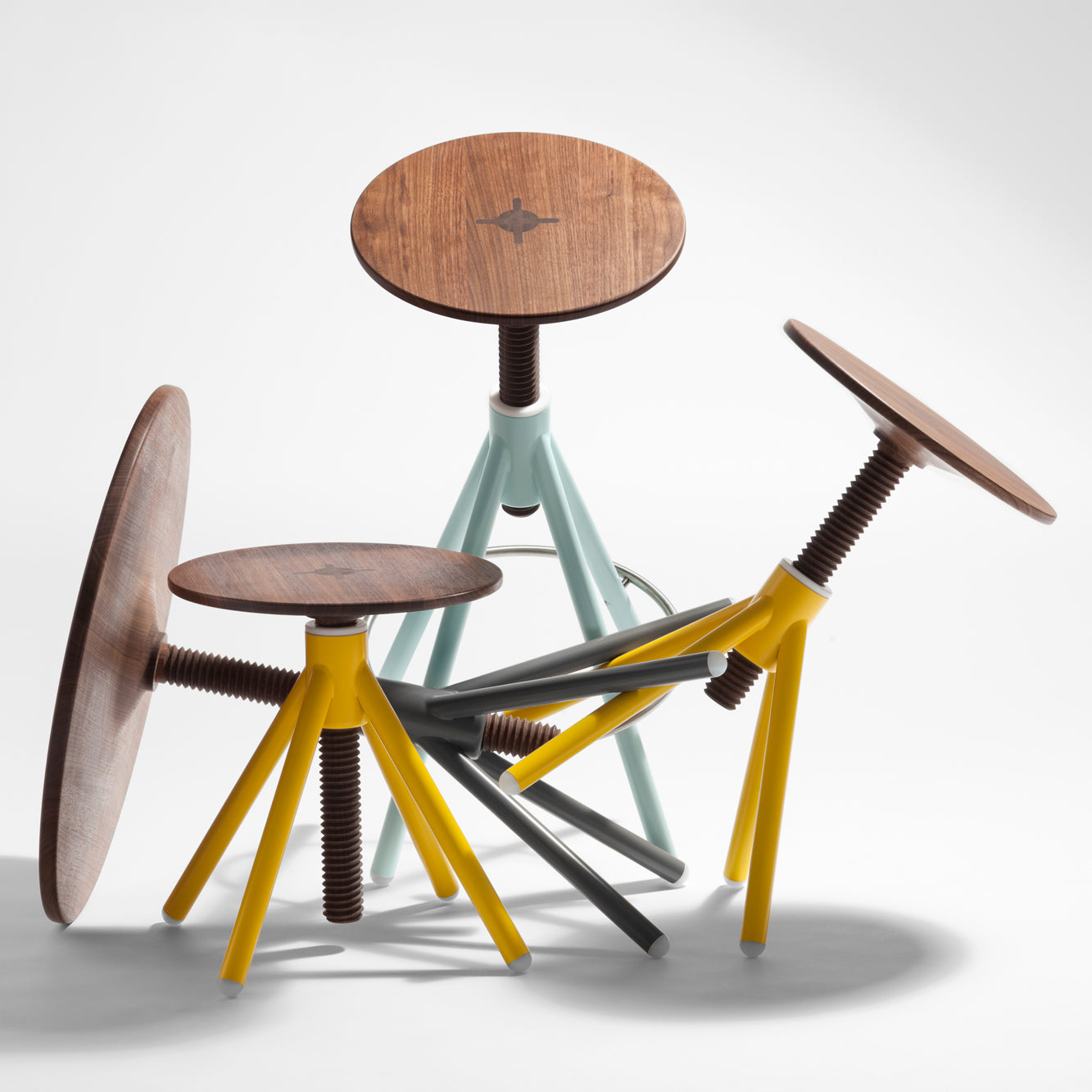 Three customizable Thread Family stools by Coordination Berlin are crafted from walnut with adjustable seats and legs in yellow, blue, and gray. Arranged on a white surface, two of the stools are positioned overturned in an overlapping manner.