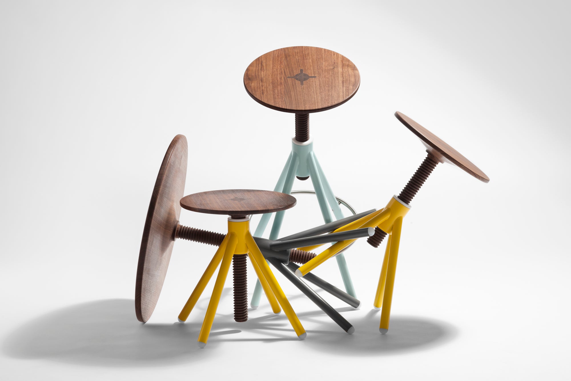 Three high stools from Coordination Berlins Thread Family feature round walnut seats and colorful metal screw bases in pastel blue, yellow, and gray. Each adjustable stool showcases a star design on its seat against a light background.