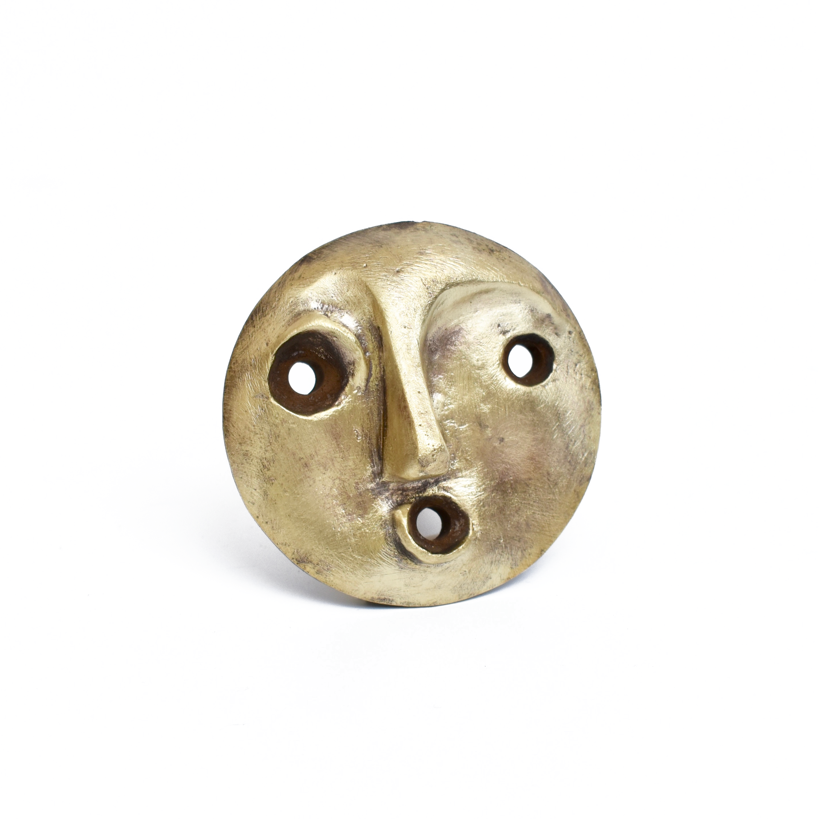 A round, metallic bottle stopper, Peonio #4685 by Werkstätte Carl Auböck, resembles a face with simple features such as a raised nose and mouth, and three holes for the eyes and mouth. Its weathered golden hue echoes pieces from the Carl Auböck workshop showcased at Triennale di Milano 1954.