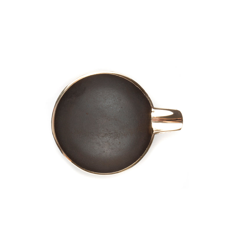 The Ashtray #4082 by Werkstätte Carl Auböck, features a shallow, round black cast iron body with a gold-colored rim and handle. Its photographed from above on a plain white background.