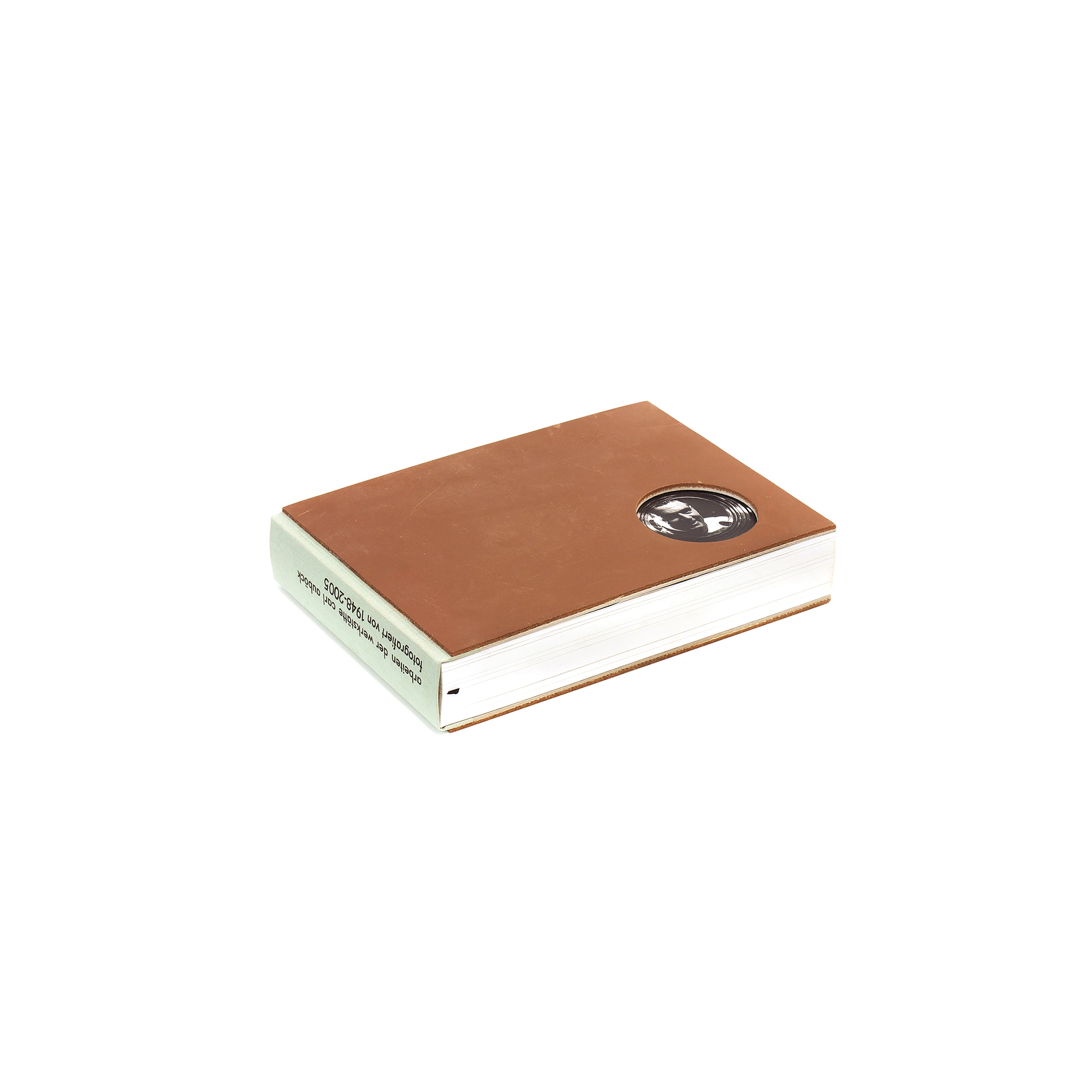 The Arbeiten der Werkstaetten Carl Auböck by Werkstätte Carl Auböck is a brown hardcover book with a minimalist black and white circular sticker on the cover, elegantly showcasing its thickness and clean lines against a plain white background.