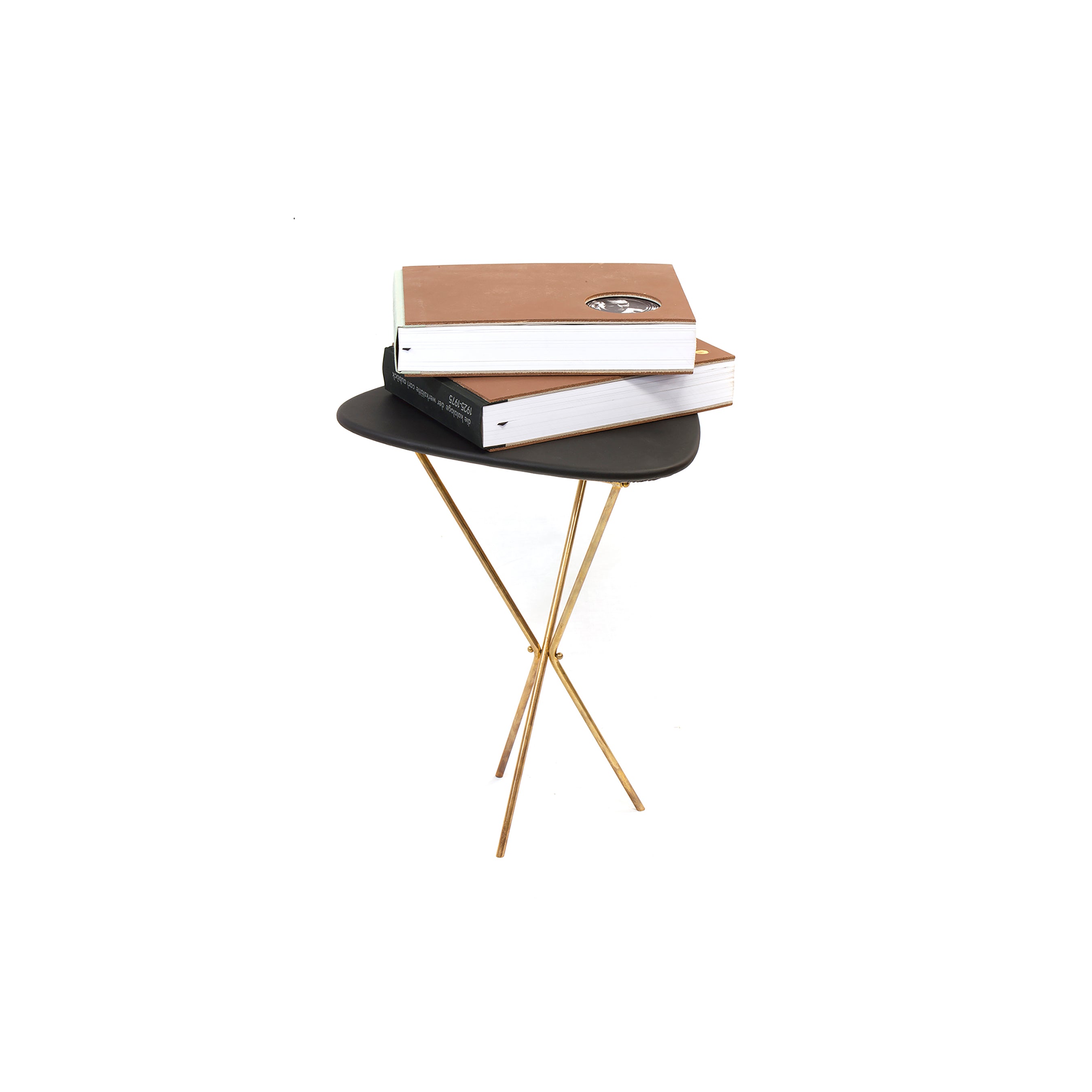 A small round black side table, reflecting Carl Auböcks minimalist style, with slender gold legs holds two large hardcover books titled Arbeiten der Werkstaetten Carl Auböck by Werkstätte Carl Auböck. The table contrasts elegantly with the substantial books against a plain white background.