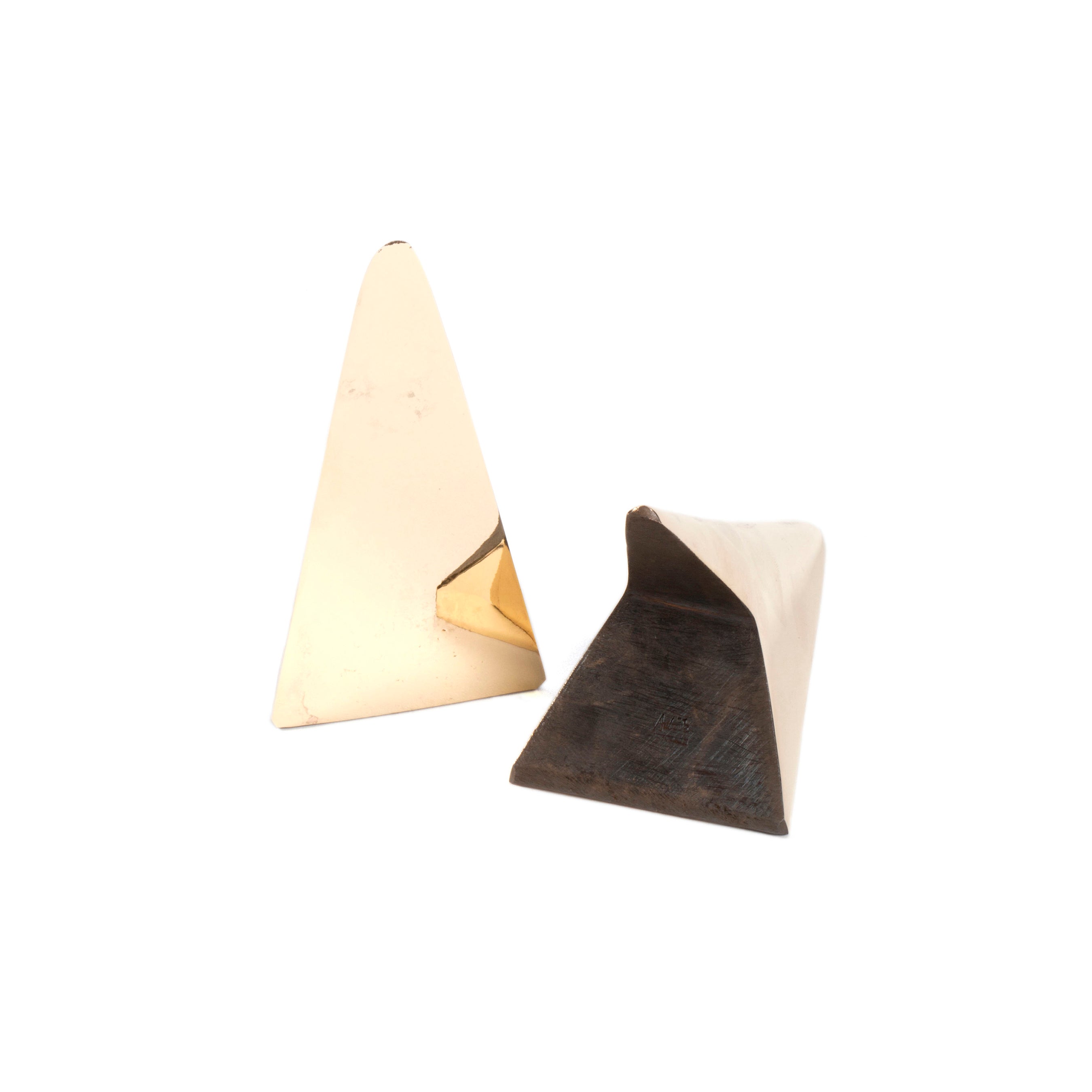 The Pair of Bookends #3846 by Werkstätte Carl Auböck feature two small, triangular gold objects on a white background. The left one stands upright, the right lies on its side, revealing a dark interior. Both have a smooth, reflective surface indicative of Wiener Bronzen artistry.