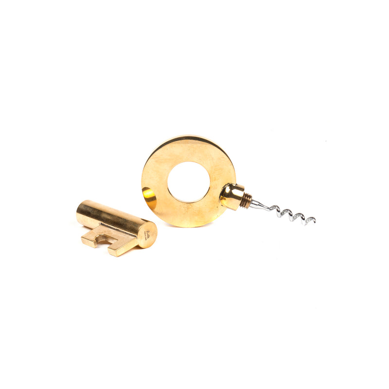 The Gold-toned two-piece set, named Corkscrew Key #5970 from Werkstätte Carl Auböck, includes a round bottle opener with a central hole and a corkscrew. Accompanied by a key-shaped opener, it showcases the timeless elegance and meticulous craftsmanship of Auböck against a white background.