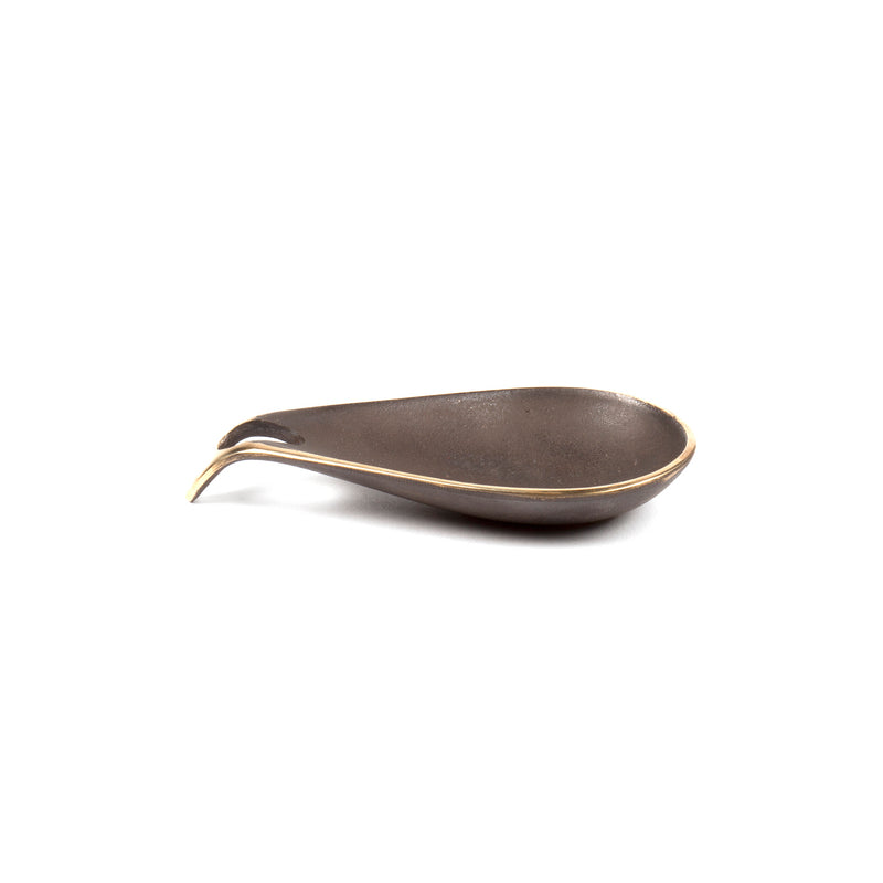 The Dish #3904 by Werkstätte Carl Auböck, with its sleek dark brown metal and gold-edged scoop and elongated pointed handle, evokes a timeless elegance reminiscent of a Viennese townhouse against a white backdrop.