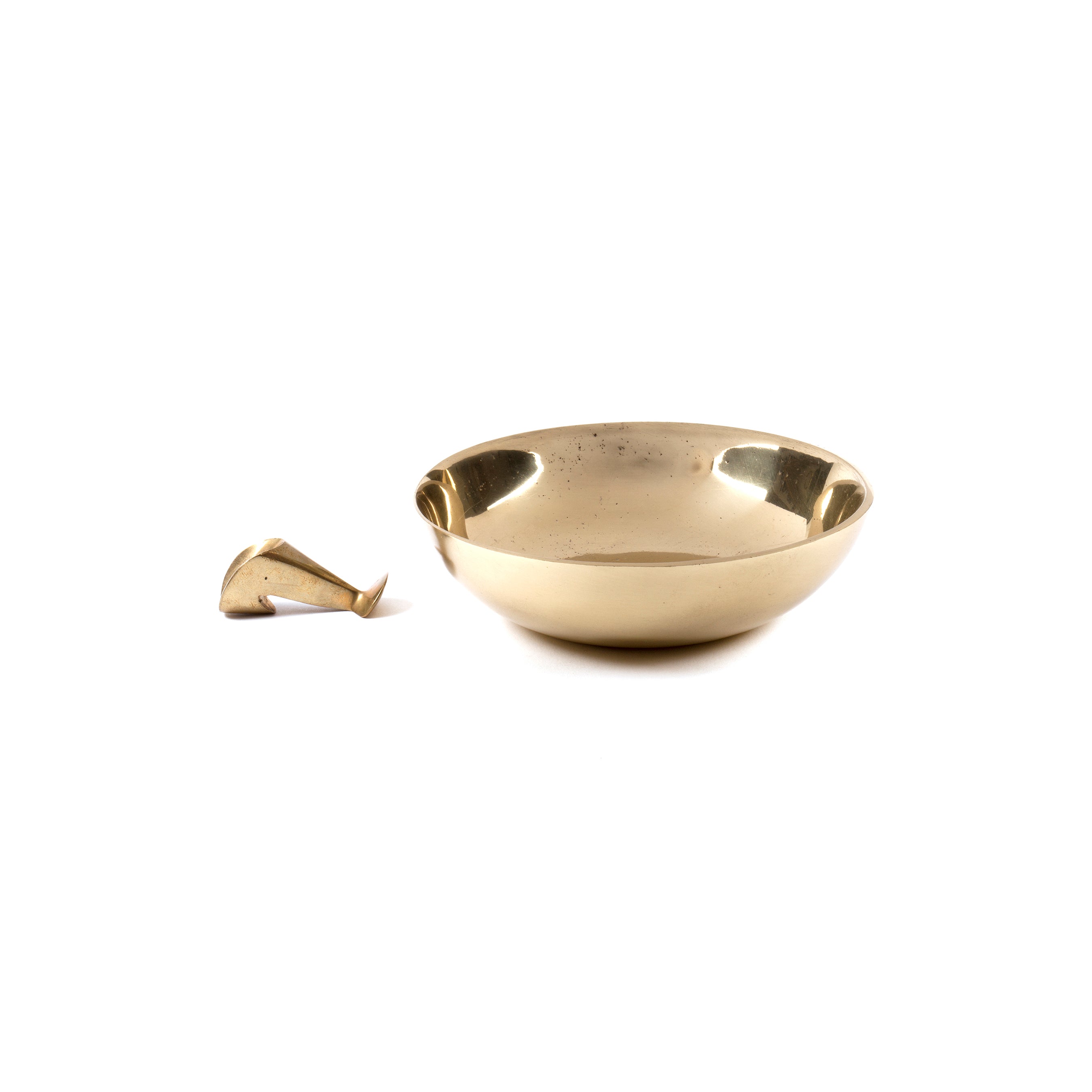 A reflective, round brass ashtray with snuffer #4053 from Werkstätte Carl Auböck sits on a plain white backdrop. To its left, a small abstract brass piece lies apart, both embodying the Wiener Bronzen style craftsmanship.