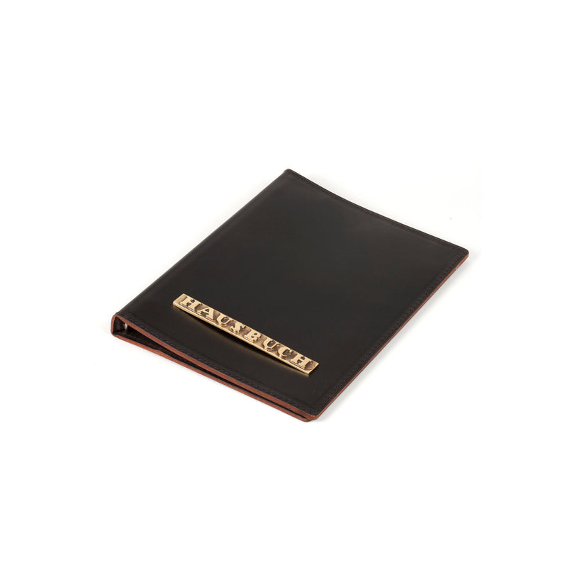 A closed black guestbook from Werkstätte Carl Auböck, featuring a sleek cover and a metal plaque embossed with TAXBUCH on the front, is angled slightly to the left against a white background.