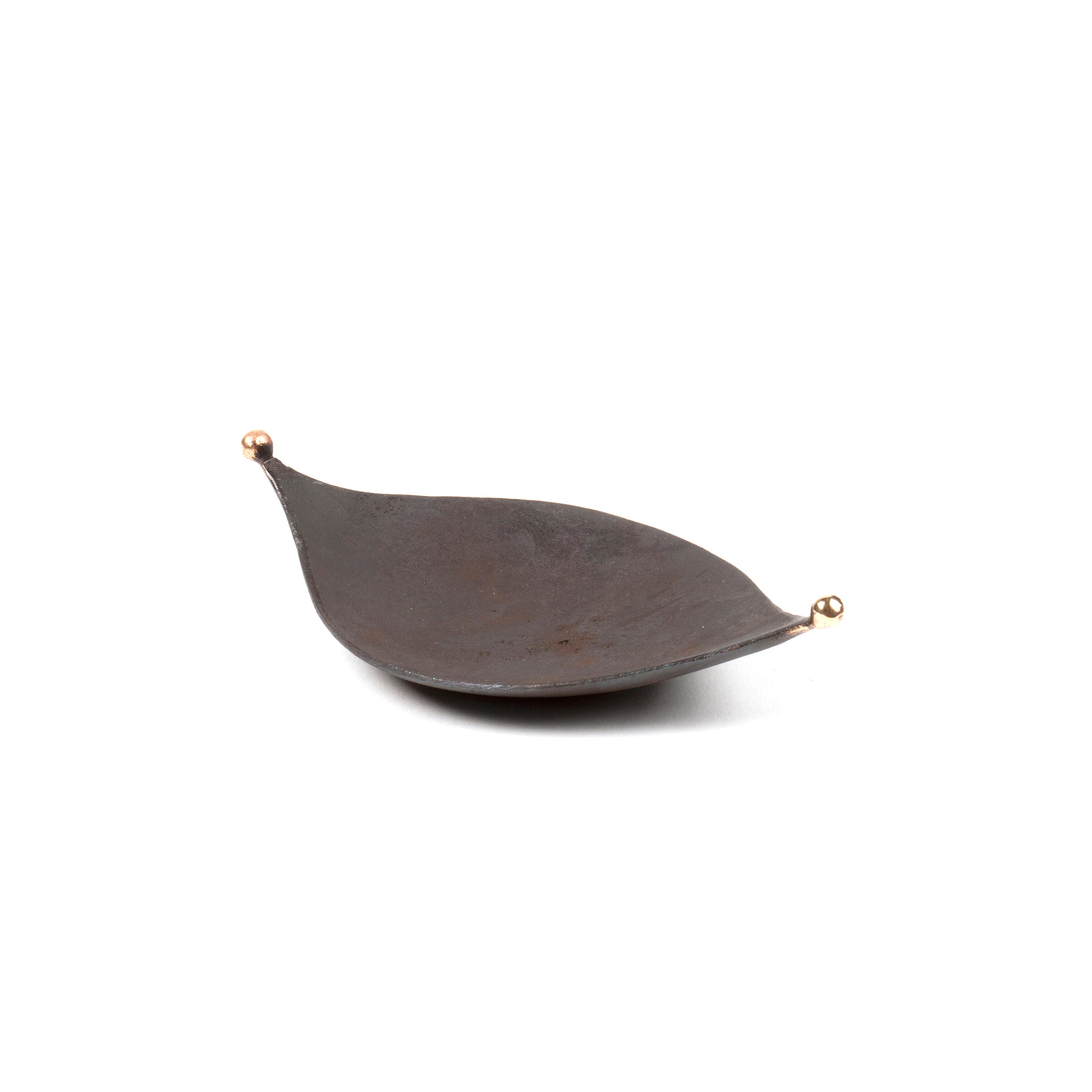 The Knopferl Tray #3431 by Werkstätte Carl Auböck is a dark, leaf-shaped metal dish with pointed ends adorned by small gold-colored spheres. Its smooth, slightly curved surface rests against a plain white background, echoing the elegance of Triennale di Milano 1954.