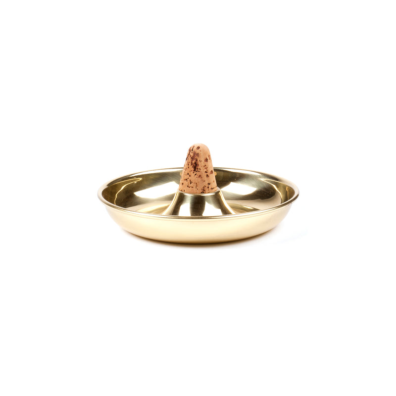 This gold-colored metal incense holder by Werkstätte Carl Auböck, called Pipe Ashtray #7202, features a central cone-shaped incense stick. Its shiny, round surface elegantly reflects light, and the brown incense cone displays a speckled pattern.