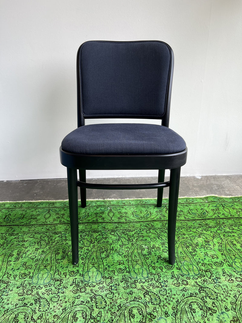 The Chair 811 by TON, on sale, showcases a Josef Hoffmann-inspired design with a curved backrest and black wooden legs. Its elegantly placed on a vibrant green patterned rug against a plain white wall and sleek gray concrete floor.