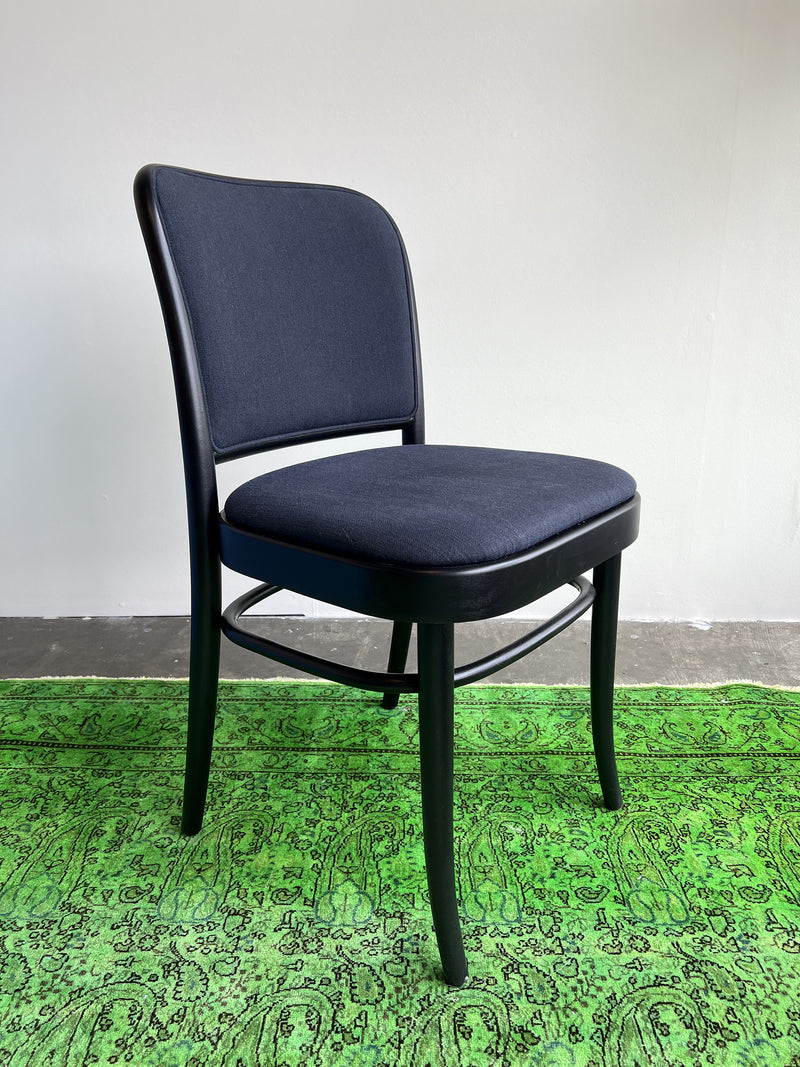 A dark navy Chair 811 by TON, featuring curved legs and a cushioned backrest in an Art Nouveau style, stands on a bright green patterned rug against a plain white wall and gray floor.