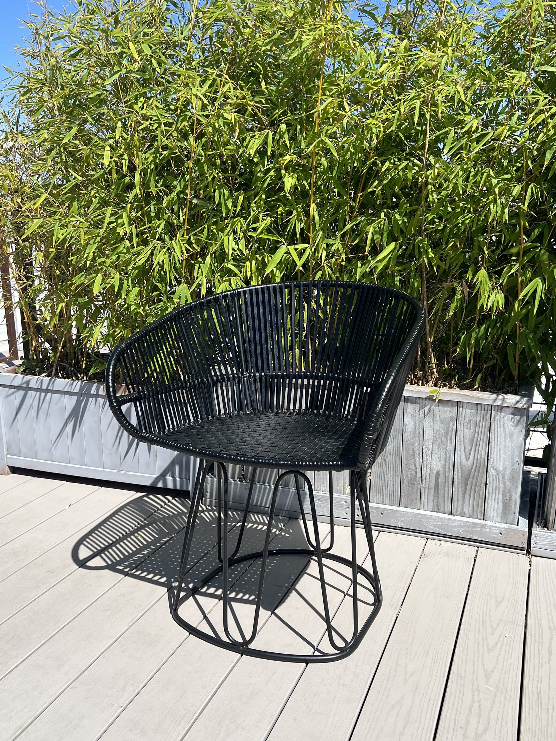 A black Circo chair from Ames, designed by Sebastian Herkner, adorns a wooden deck. Behind it, tall bamboo plants in a planter flourish under the clear blue sky.