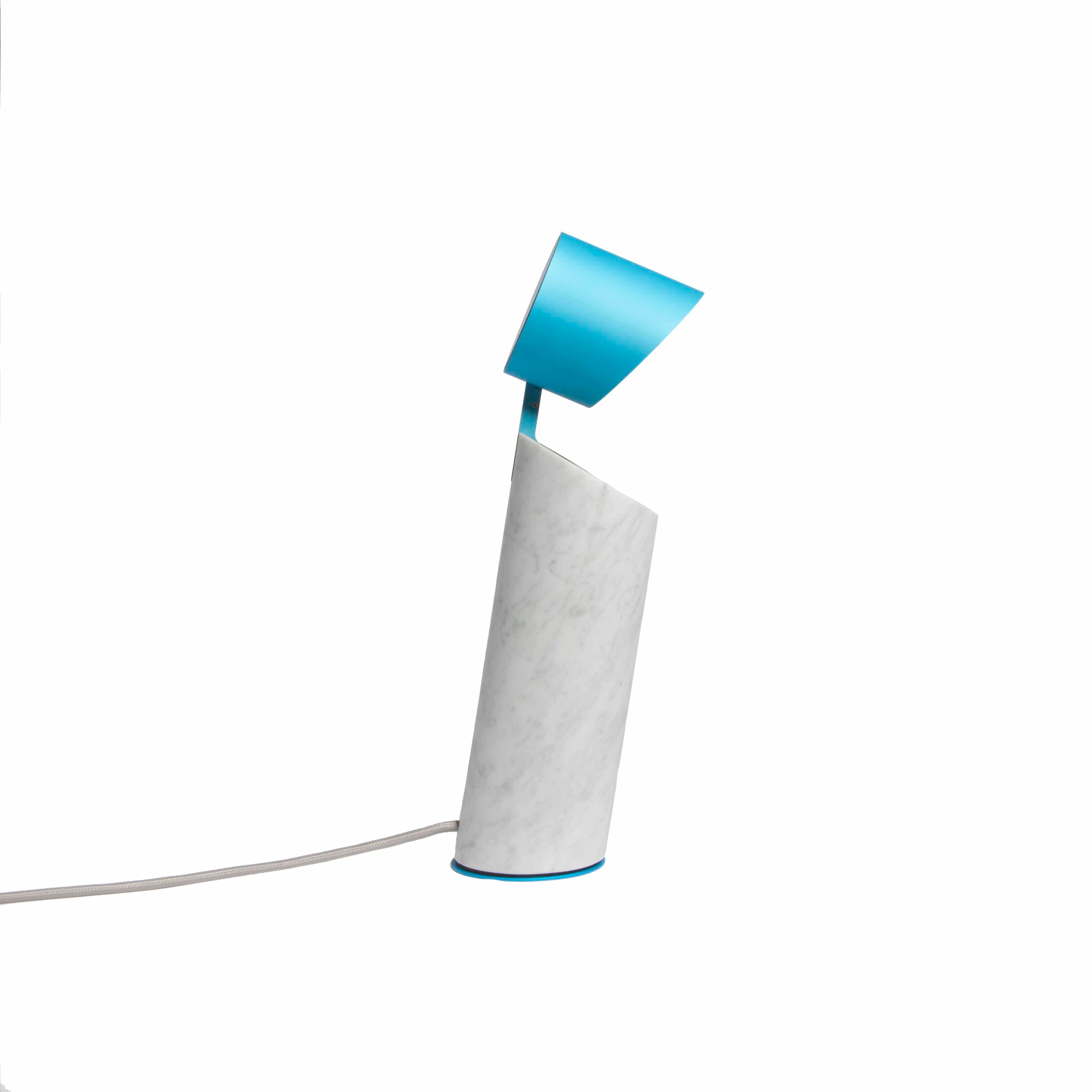 The Italic Light by Coordination Berlin is a modern lamp with a sculptural cylindrical white marble base and a blue angular shade, featuring an LED ring for efficient lighting and a power cord from the base, set against a plain white background.