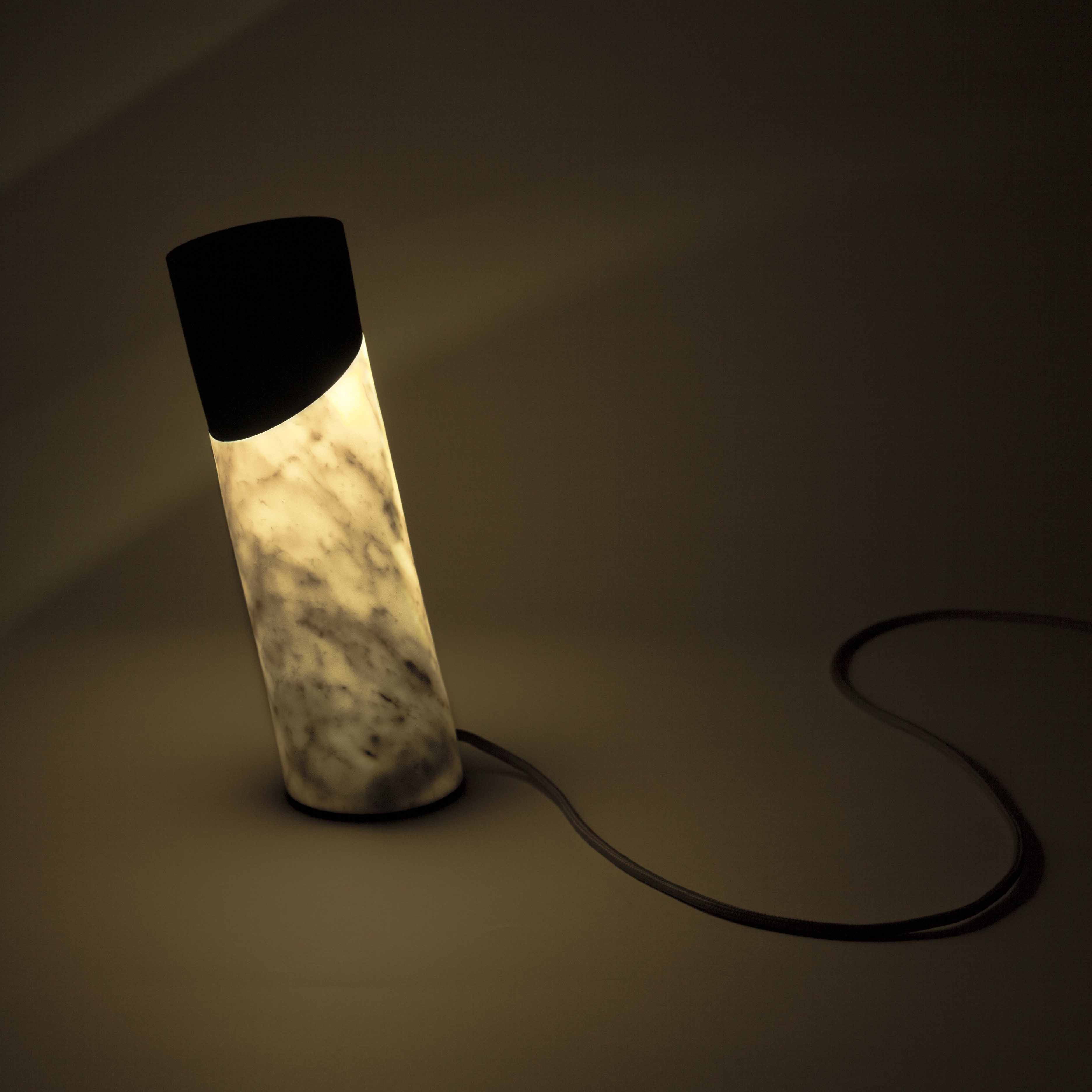 The Italic Light by Coordination Berlin is a modern, cylindrical lamp featuring a marbled base and sculptural side light. It leans slightly on a smooth surface, with a power cord casting shadows, while an elegant LED ring emits a captivating warm glow in the dimly lit setting.