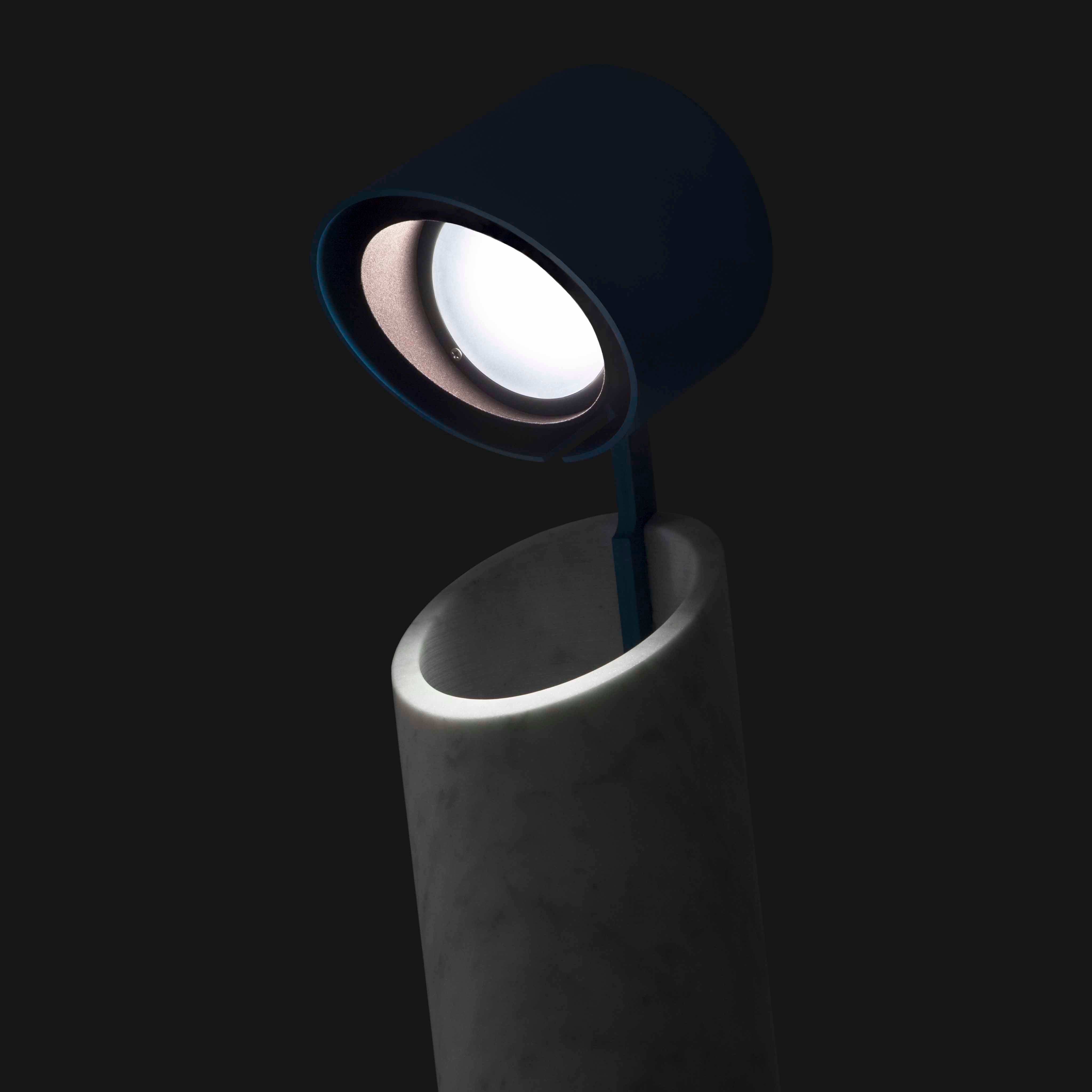 The Italic Light by Coordination Berlin is a modern desk lamp with a cylindrical marble base and dark rounded lampshade. Its sculptural design includes a minimalistic arm extending to an LED ring, casting an elegant glow against a black background.