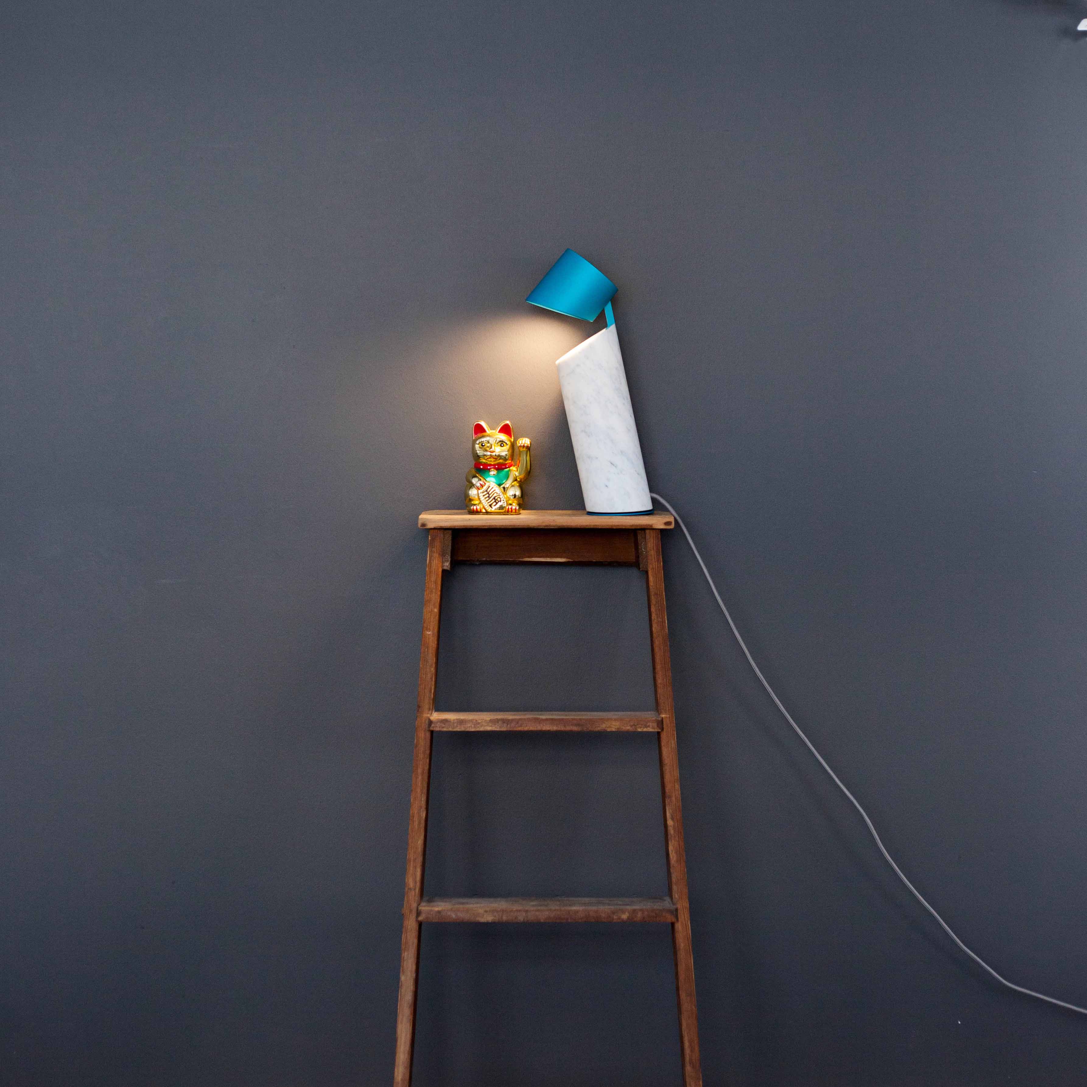 The Italic Light by Coordination Berlin, featuring a blue shade, illuminates a ceramic waving cat atop the wooden ladder against a gray wall, enhanced by a sculptural side light while a power cable runs down the ladder.