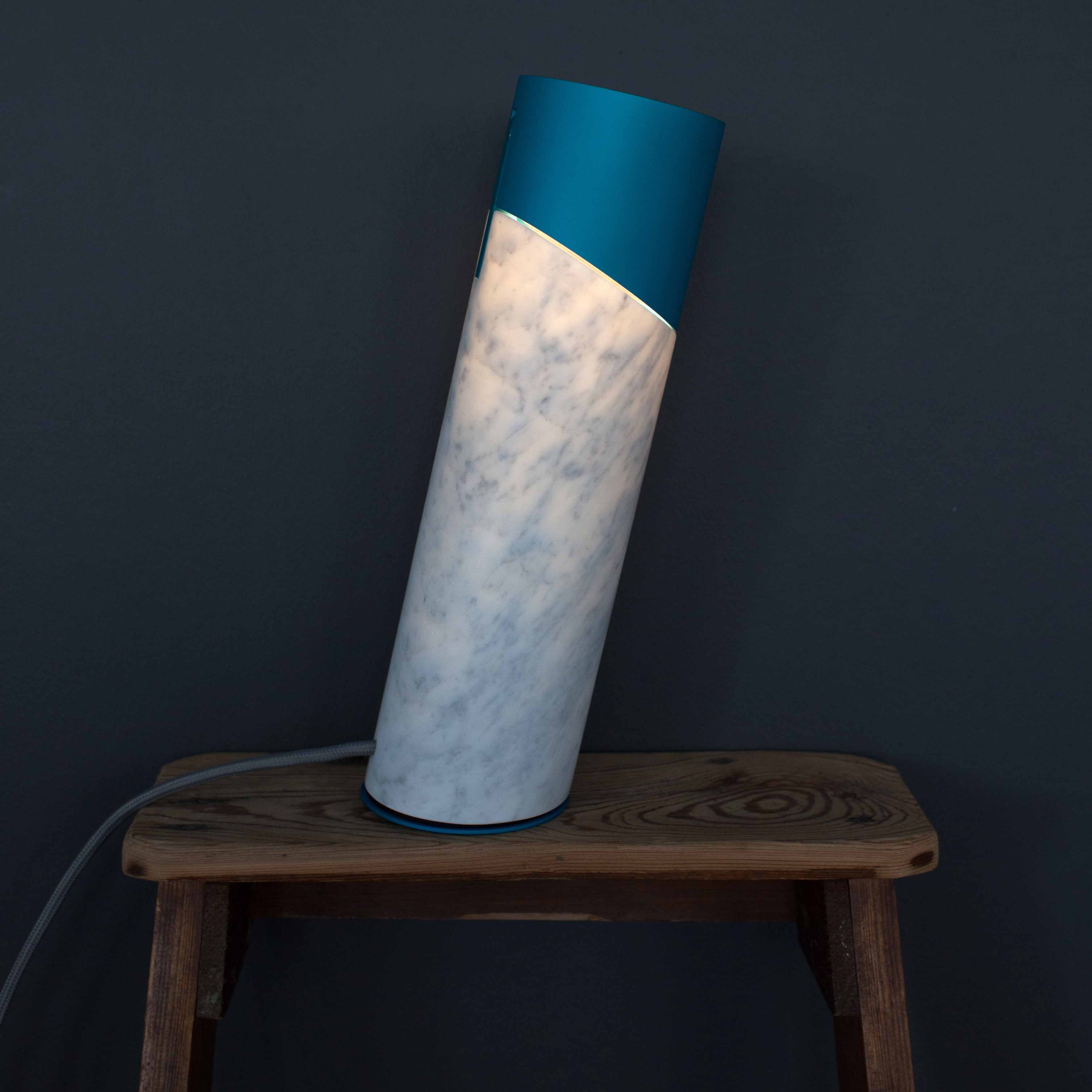 The Italic Light by Coordination Berlin is a cylindrical sculptural side lamp with a marble-like texture and blue top, sitting tilted on a wooden stool against a dark gray background. Enhanced by an LED ring, it casts a soft glow with the cord elegantly extending from the base.