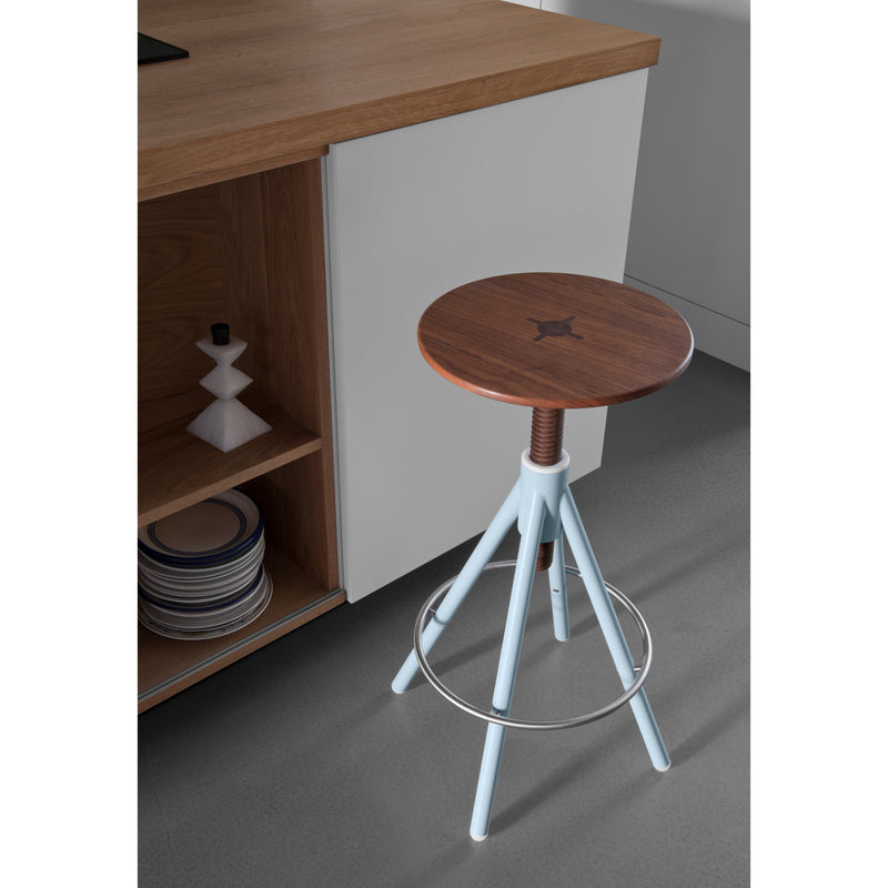 A Coordination Berlin Thread Family High Stool with a wooden top, circular metal footrest, and light blue legs stands next to a wooden kitchen cabinet. The cabinet has an open shelf containing stacked plates and a geometric ornament on a gray floor.