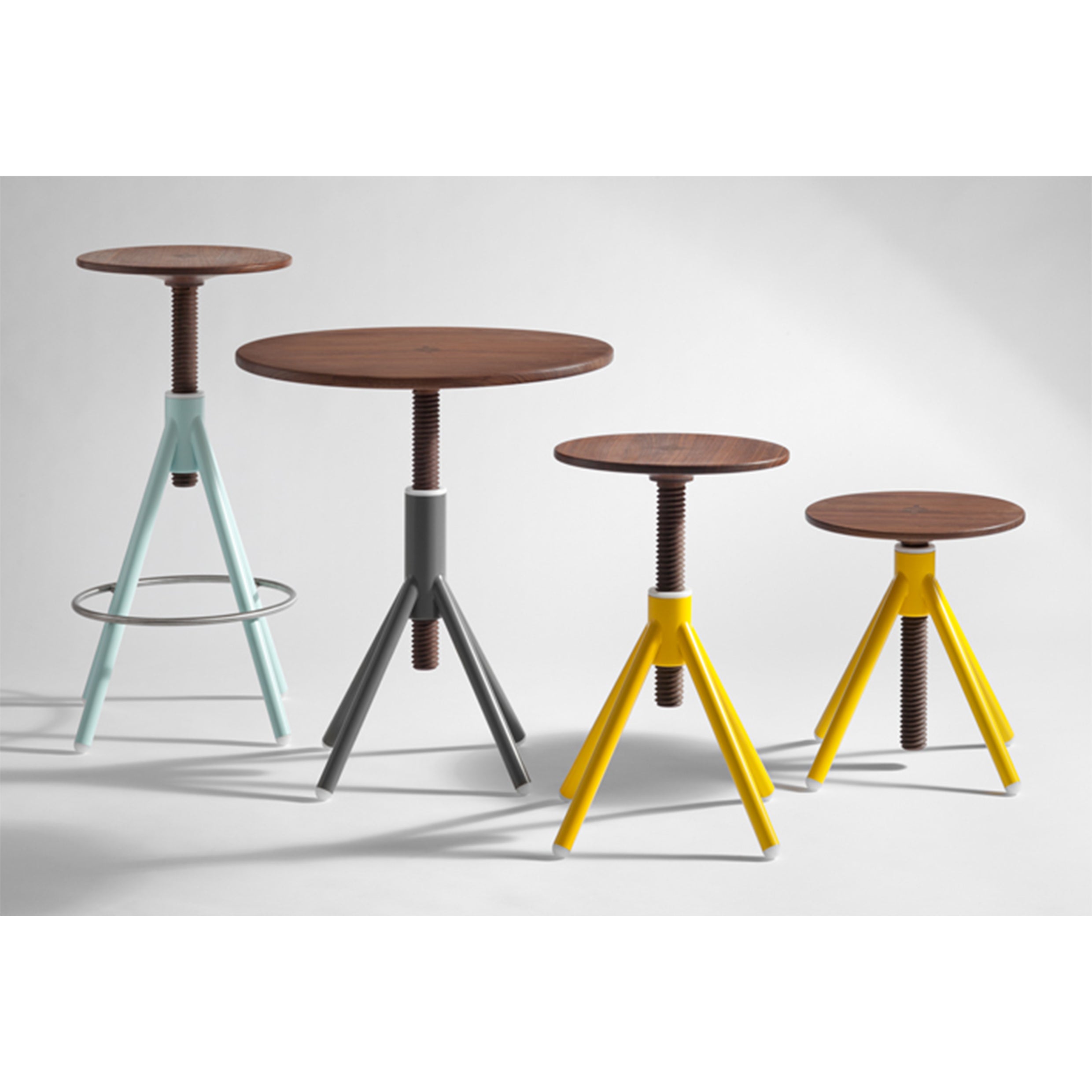The Thread Family high stools by Coordination Berlin feature adjustable heights, circular walnut seats, and metal tripod legs in varied colors: light blue with a footrest, gray, and two yellow. They are arranged left to right from tallest to shortest.