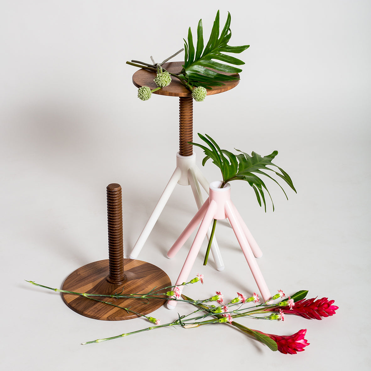 Three Thread Family - Stools by Coordination Berlin, crafted from walnut wood, stand on a white surface with green foliage on top. One stool is upside down. Red and pink flowers are scattered around, enhancing the minimalist setup with vibrant colors.