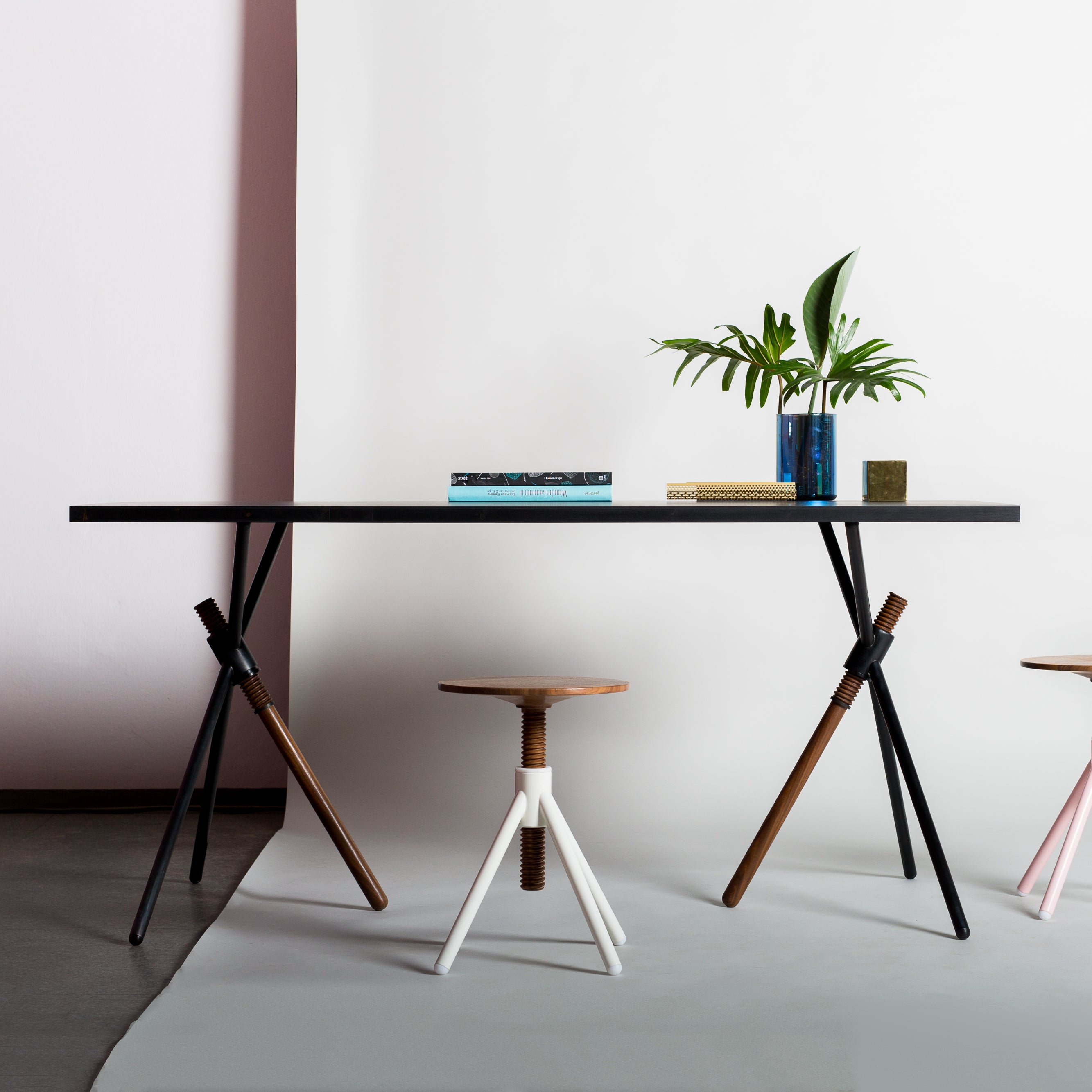 The Thread X by Coordination Berlin is a contemporary table with adjustable angled legs, two stools—one with sleek white legs and the other with warm wooden ones—set against a soft pink and white wall. The tabletop displays books and a lush potted plant.