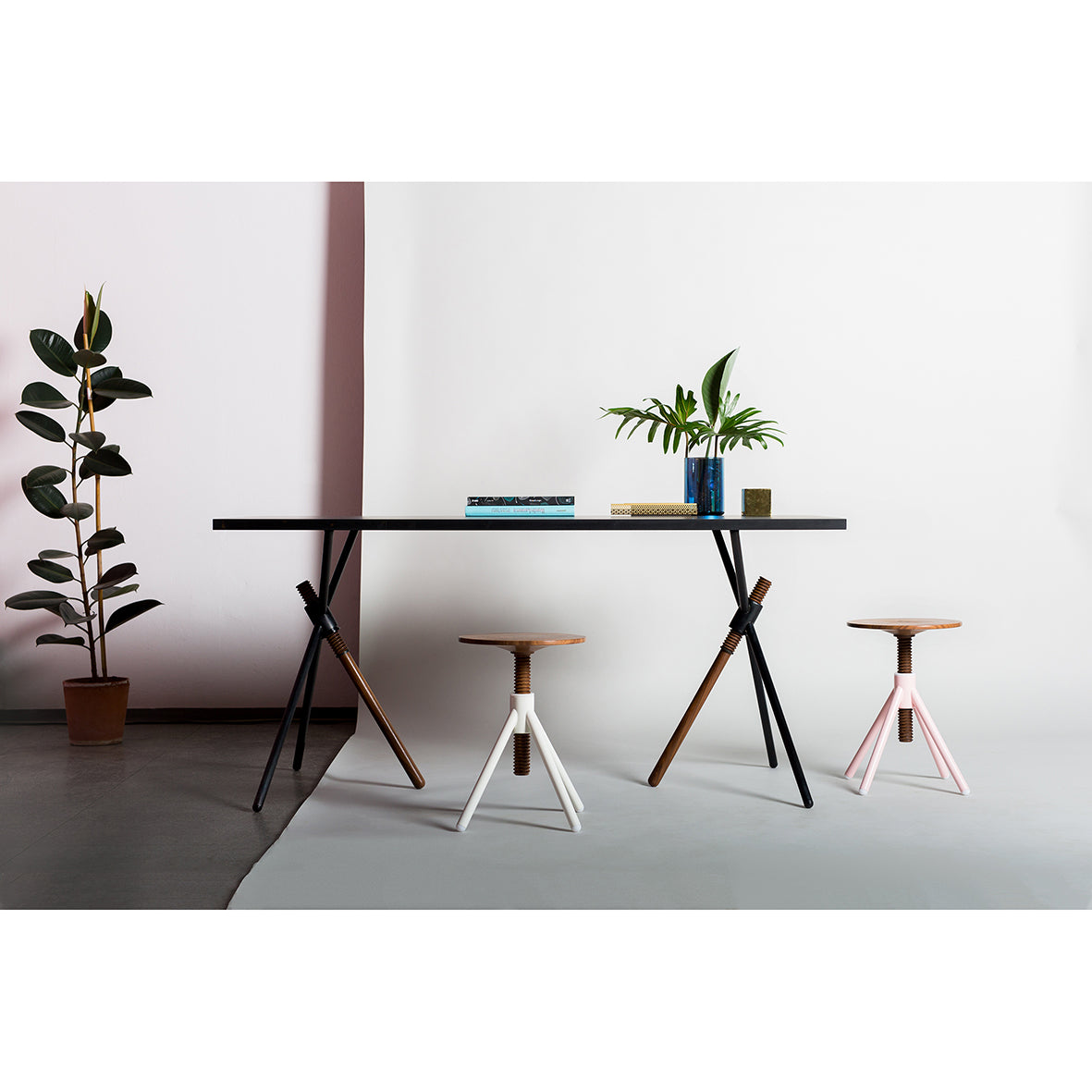 The stylish Thread X by Coordination Berlin features a modern table with a book and green plant vase, flanked by three unique stools with angled legs in black, white, and pink. A potted plant on the left completes the look against a chic white and pink two-tone wall.