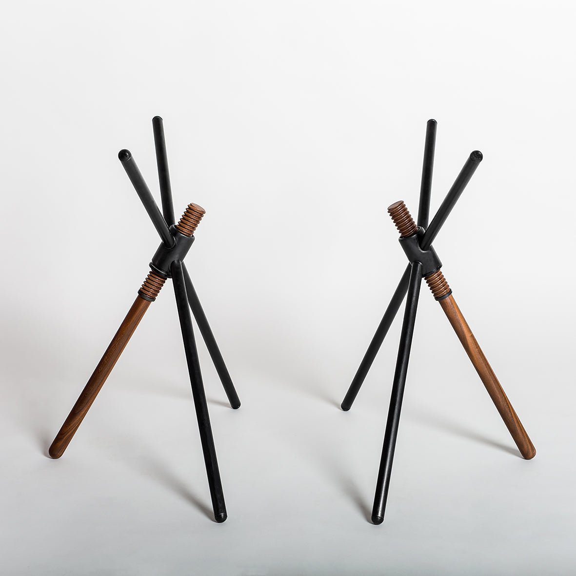 The Thread X tripods by Coordination Berlin feature a contemporary design with intersecting black rods and one wooden leg, creating a stylish geometric look reminiscent of height-adjustable furniture against a white background.