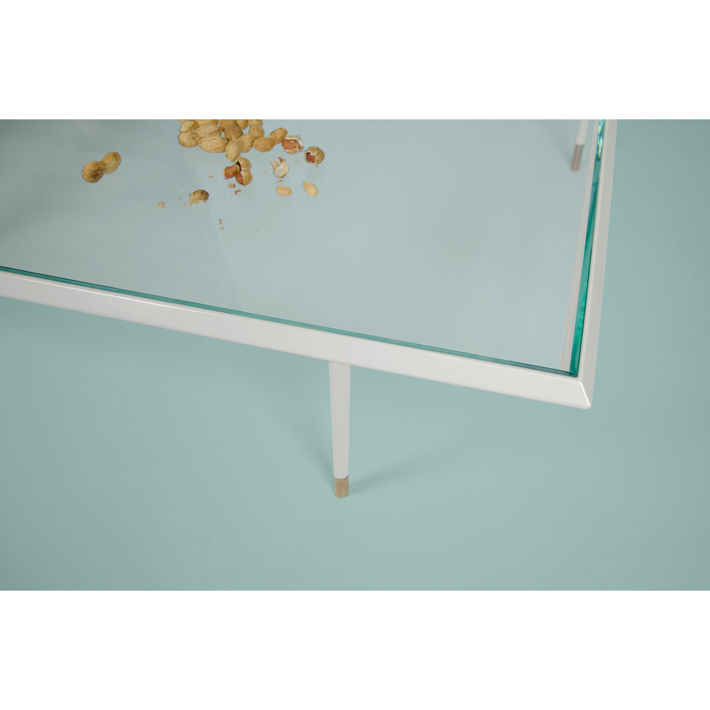 A Brutalesque Coffee Table by Coordination Berlin, featuring a glass top and white legs, sprinkled with peanut shells and crumbs. The light blue background enhances the contemporary feel of the scene.