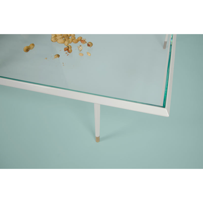 A Brutalesque Coffee Table by Coordination Berlin, featuring a glass top and white legs, sprinkled with peanut shells and crumbs. The light blue background enhances the contemporary feel of the scene.