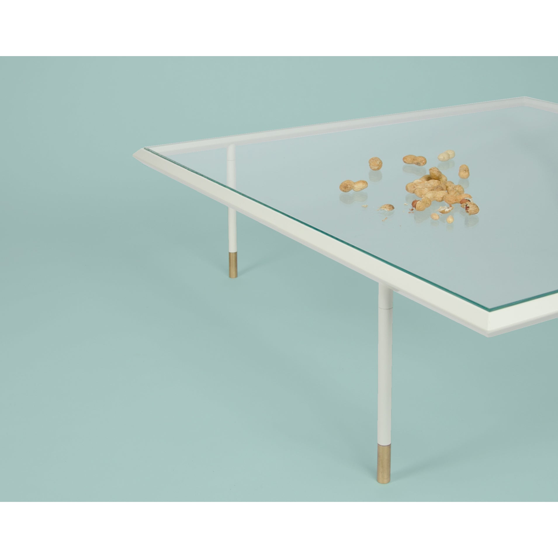 The Coordination Berlin Brutalesque Coffee Table features a glass top, white legs with brass-colored tips inspired by Brutalist Architecture, and is set against a pale blue background. A small pile of unshelled peanuts is scattered on its surface, adding an edgy touch.